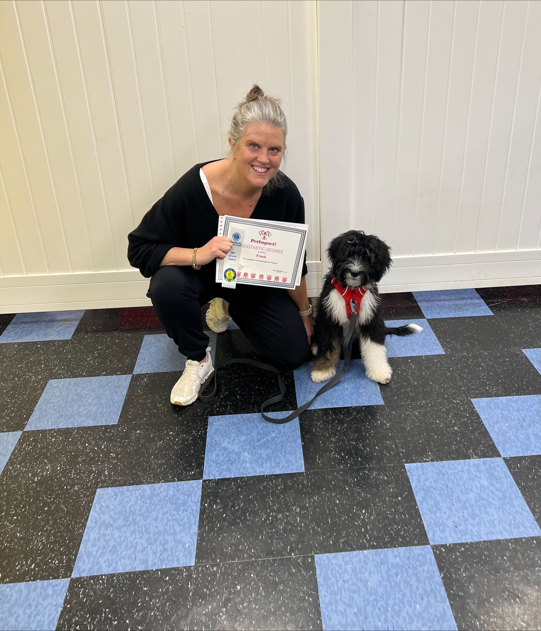 Woman kneeling with dog, holding certificate. The dog wears a red collar and is black and white. Tile floor.