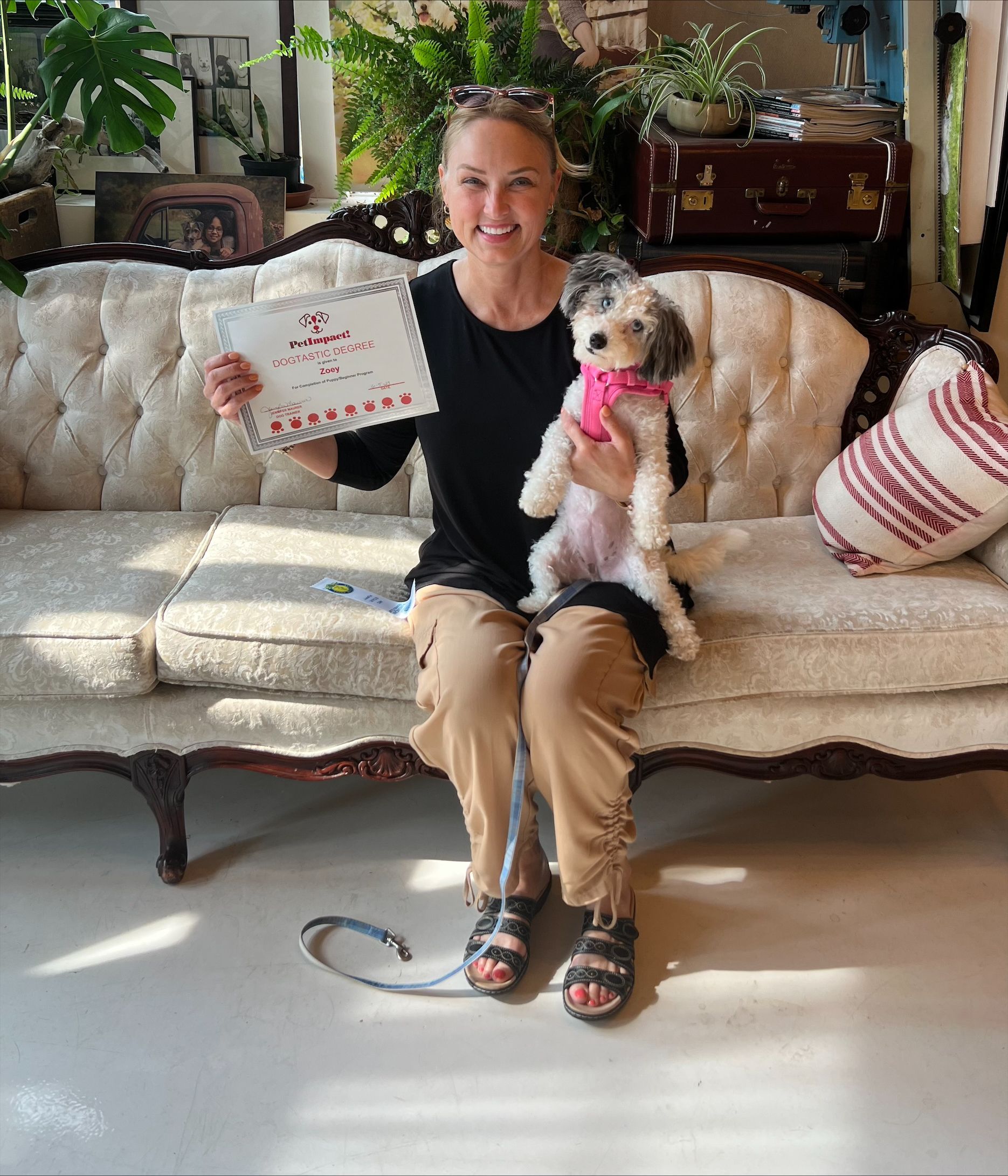 Woman on a sofa holding a dog and a card. She smiles; the dog wears a pink collar. The room is well-lit with plants.