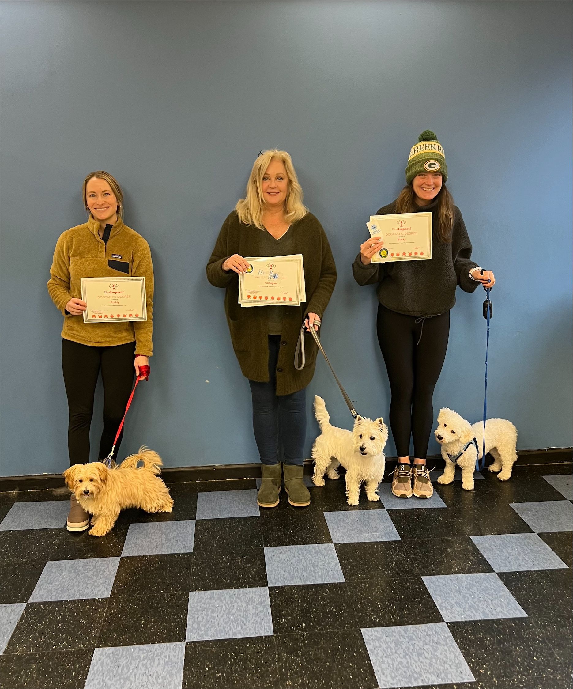 Three people with dogs pose with signs. The setting is indoors, with blue wall. Dogs are small and fluffy.