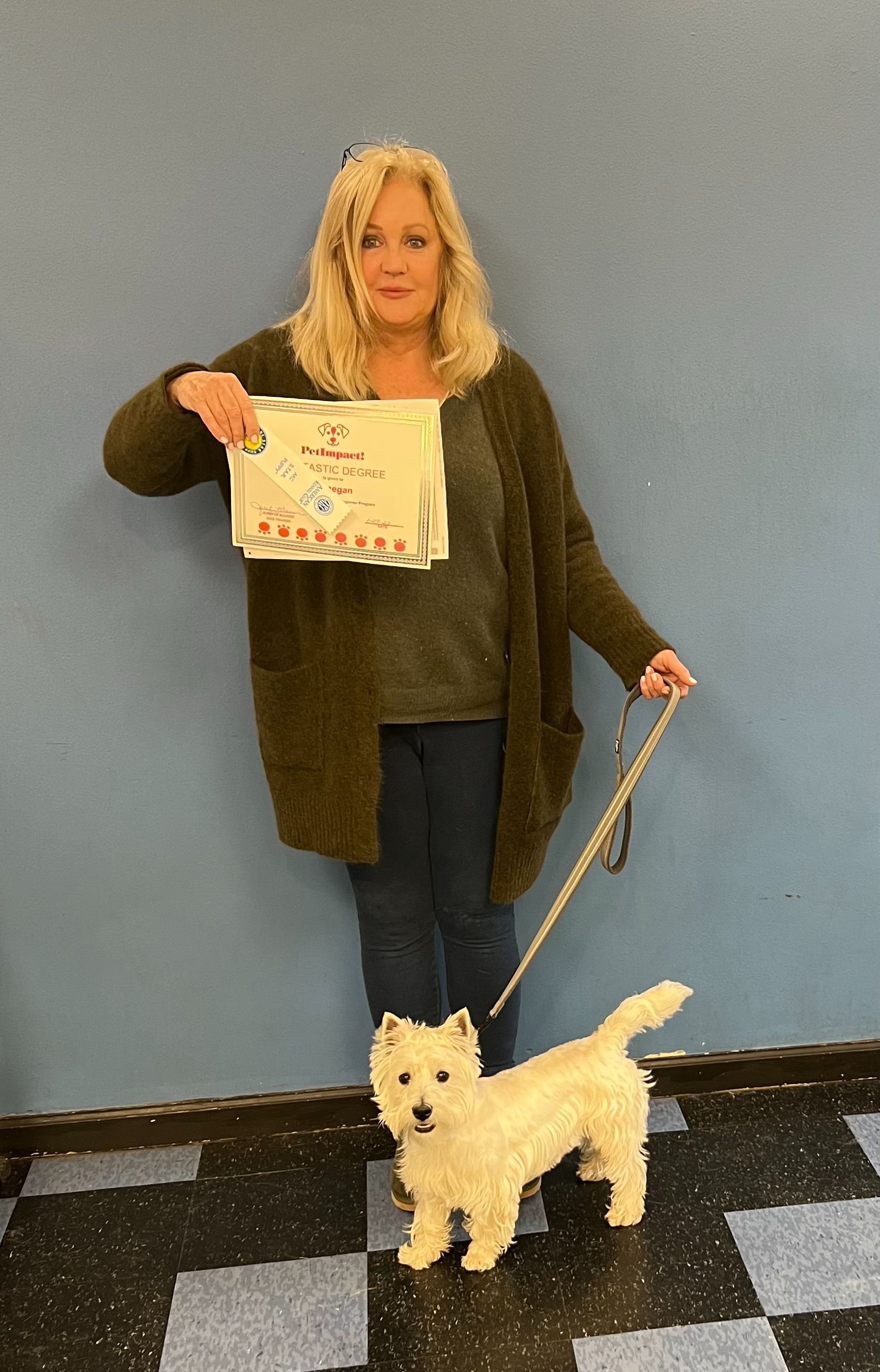 Woman holding a certificate, standing with a white dog on a leash in front of a blue wall.
