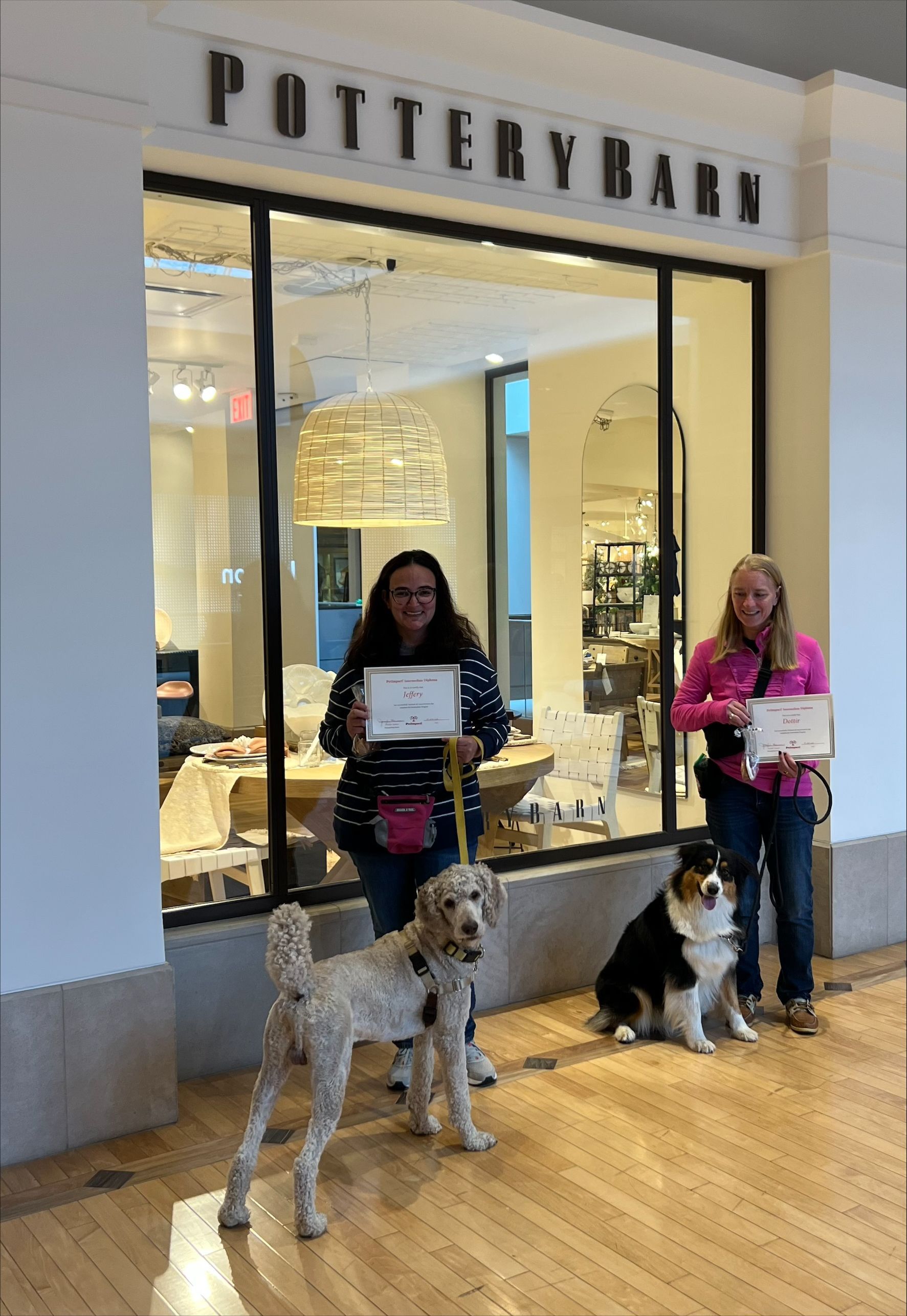 Two women with dogs in front of a Pottery Barn store, holding certificates.