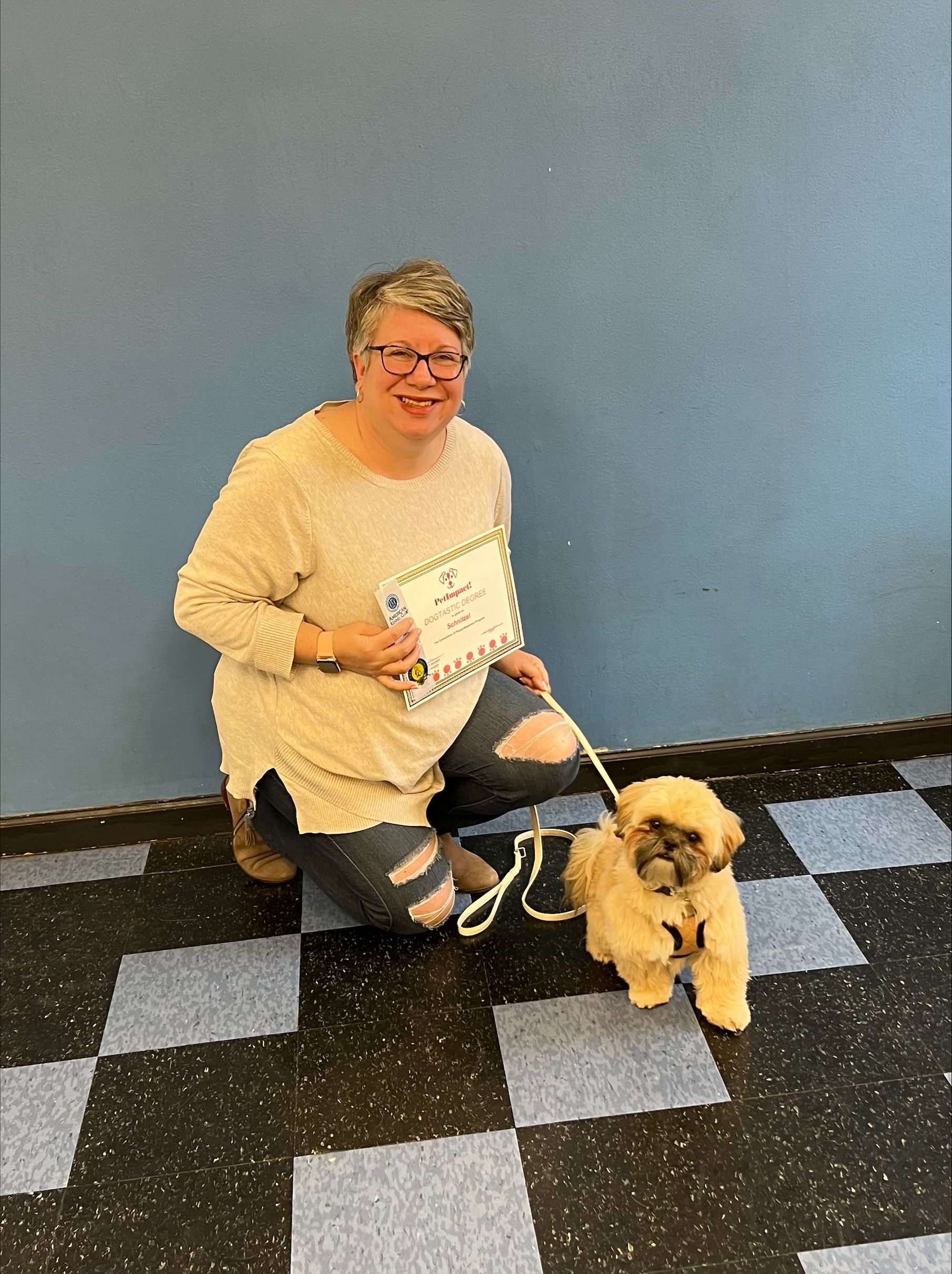 Woman kneeling beside a small, tan dog on a leash, holding a certificate. Blue wall, checkered floor.