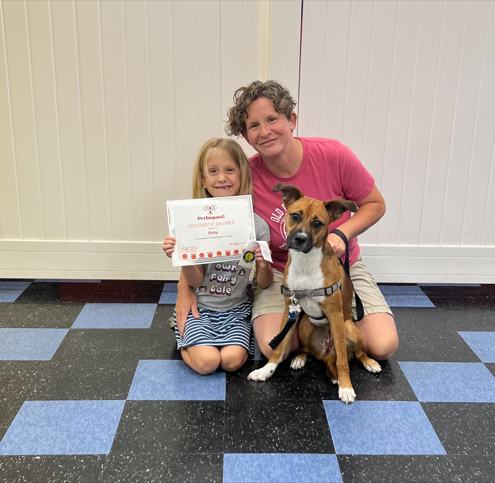 Girl and woman with dog, holding certificate. They are in a room with a patterned floor.