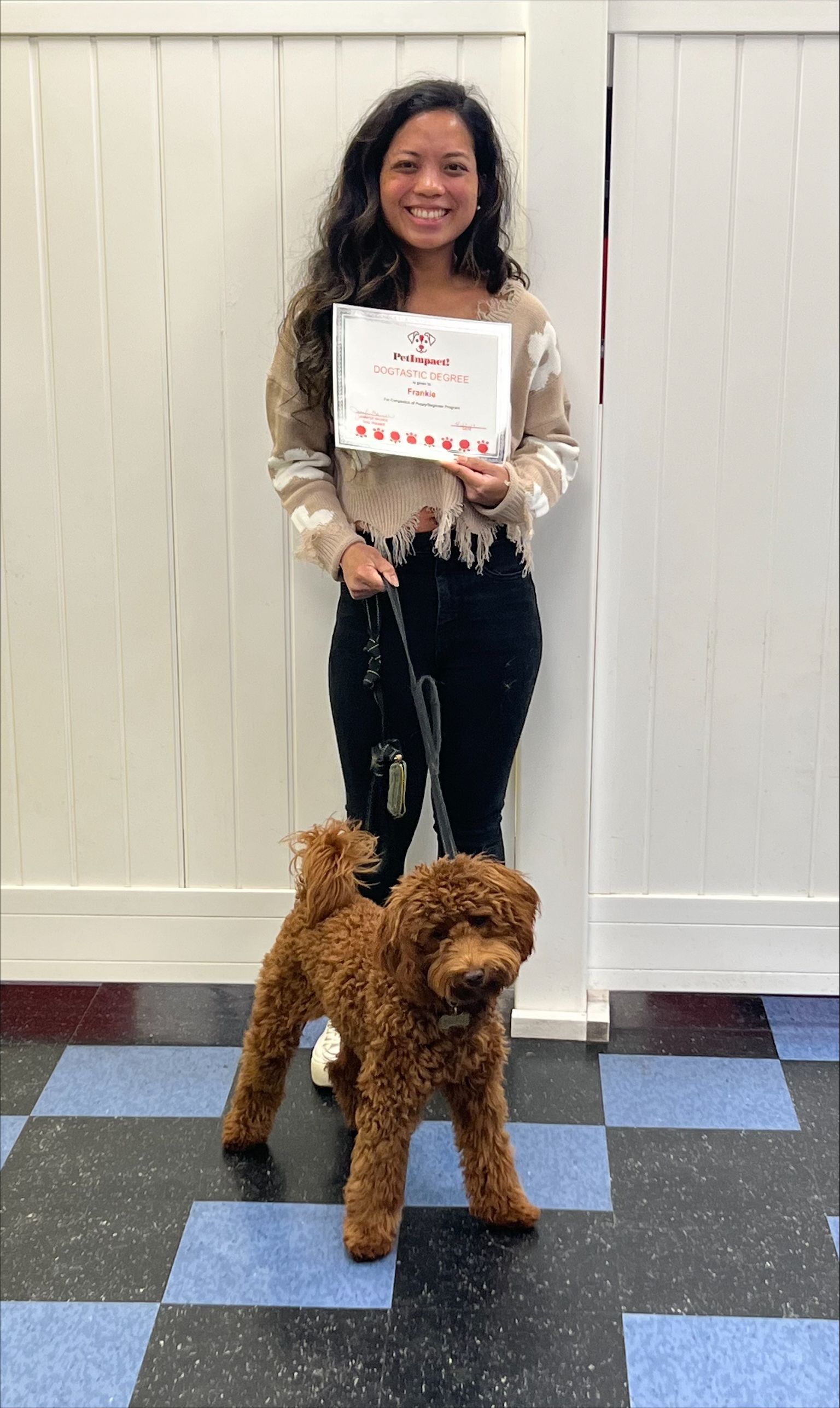 Woman holding certificate, smiling, standing with brown dog on leash. They're indoors by a checkered floor.