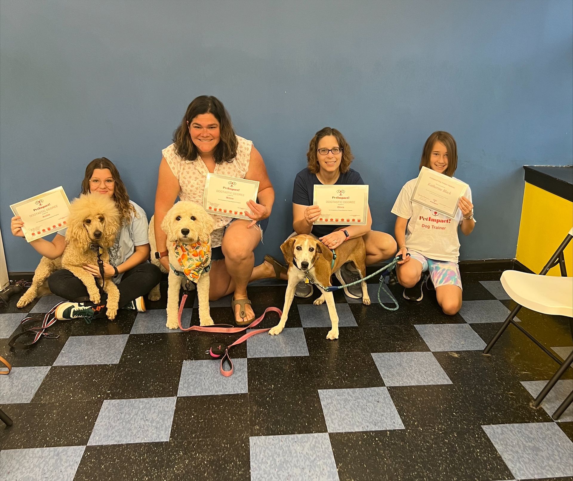 Four people and three dogs pose with certificates. They kneel on checkered floor against a blue wall.