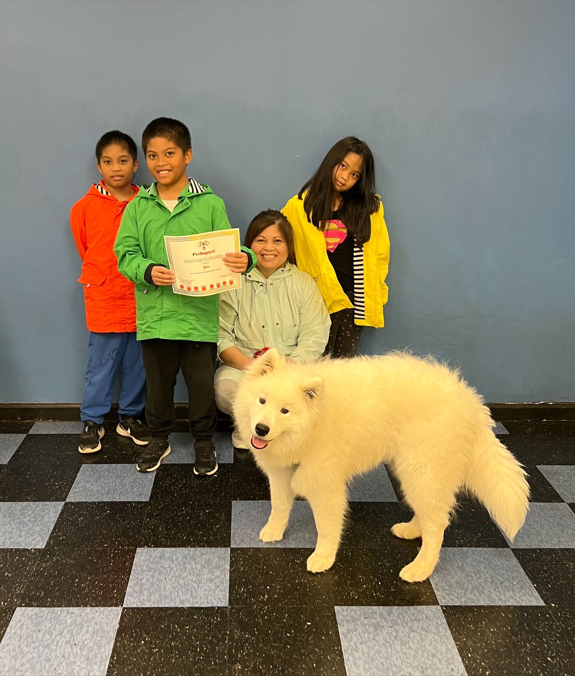 Group of children and a large white dog posing with a certificate in front of a blue wall.