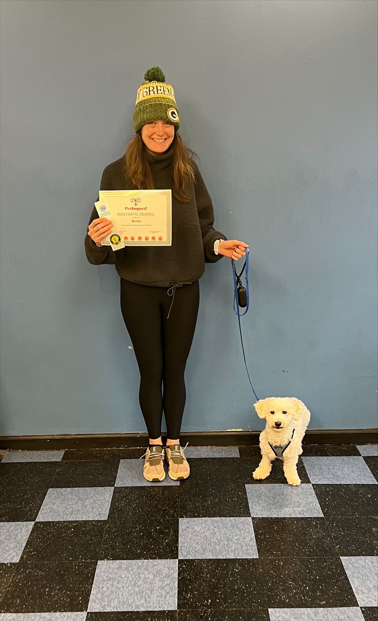 Woman in beanie and dog pose with a certificate in front of a blue wall.
