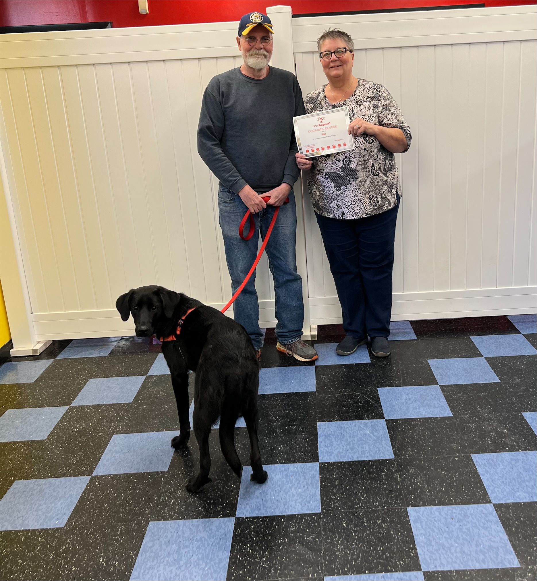 A couple with a black dog on a leash pose inside. The woman holds a certificate.