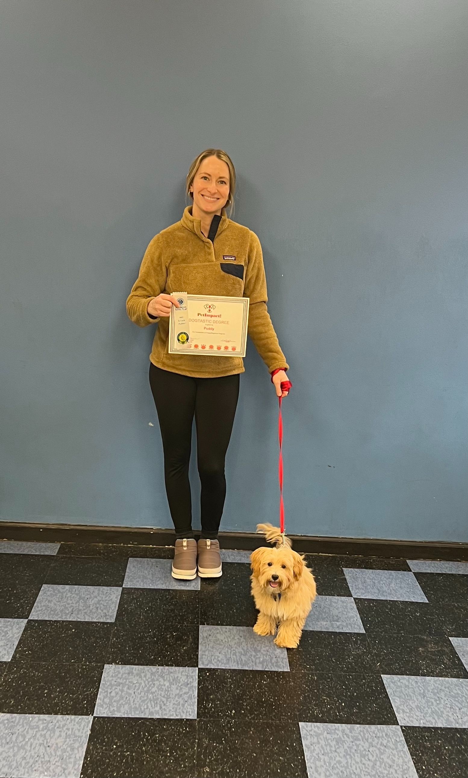 Woman smiles, holds dog's leash and a certificate. Dog is tan, leash is red. They stand by a blue wall and checkered floor.