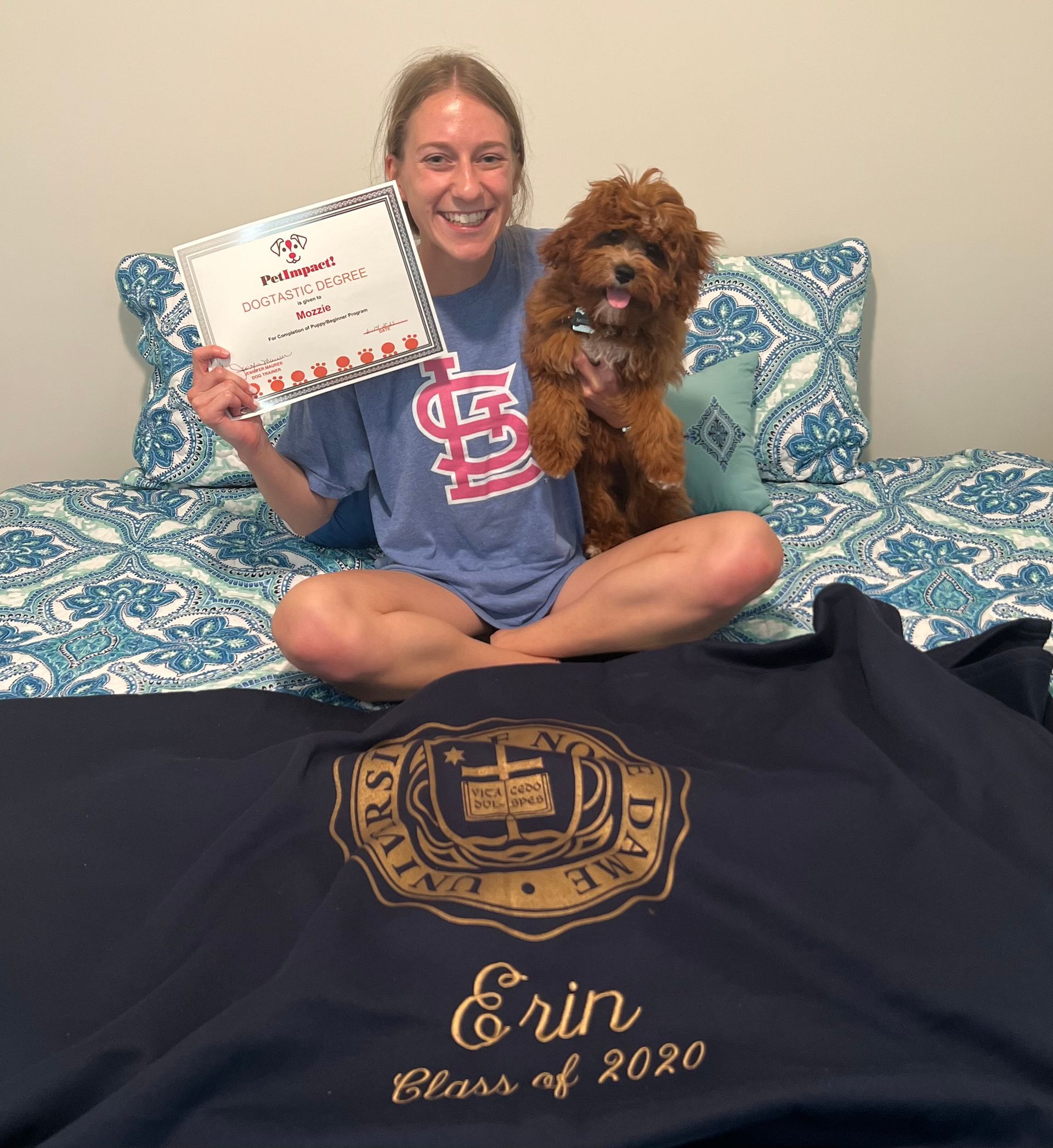 Woman with dog celebrates graduation, holding a diploma, sitting on a bed with a sweatshirt.