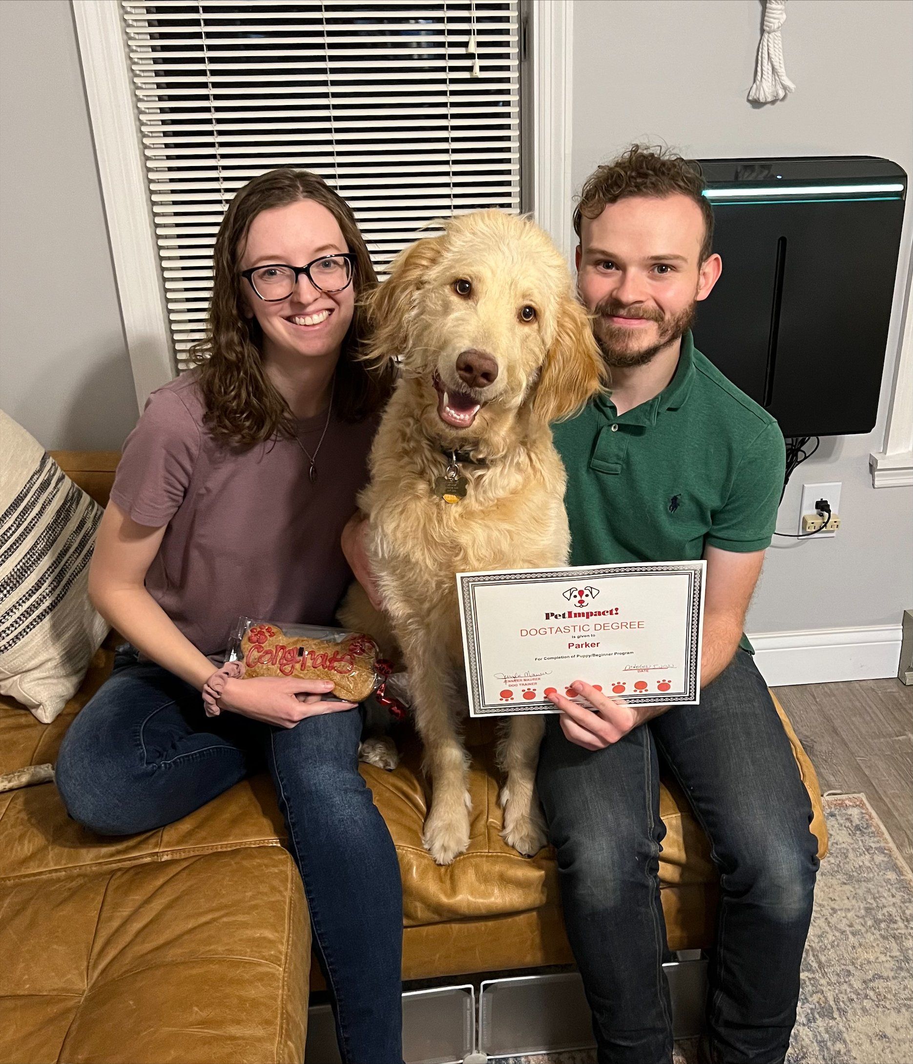 A couple and their golden doodle dog sit on a couch, holding a framed certificate.