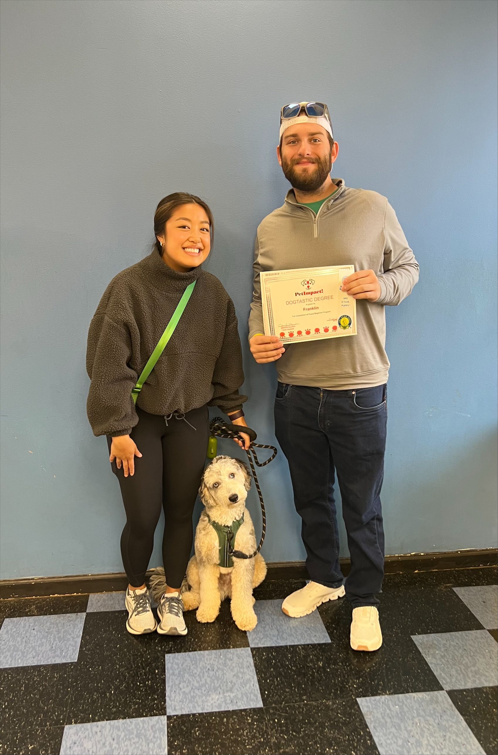 Woman, man, and service dog, smiling, holding a certificate, standing by a blue wall.