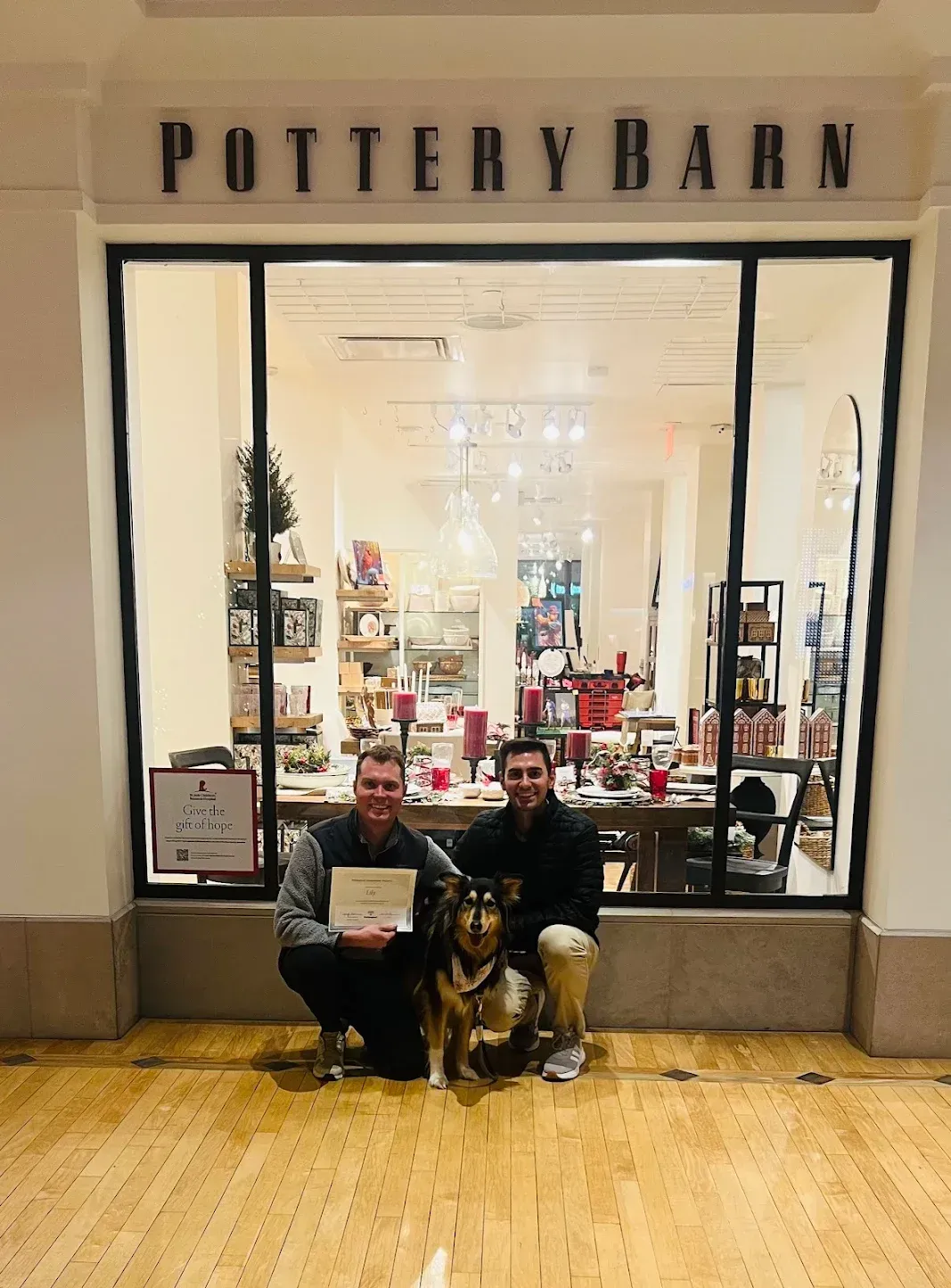 Two men and dog in front of a Pottery Barn store. One man holds a paper, all are smiling.