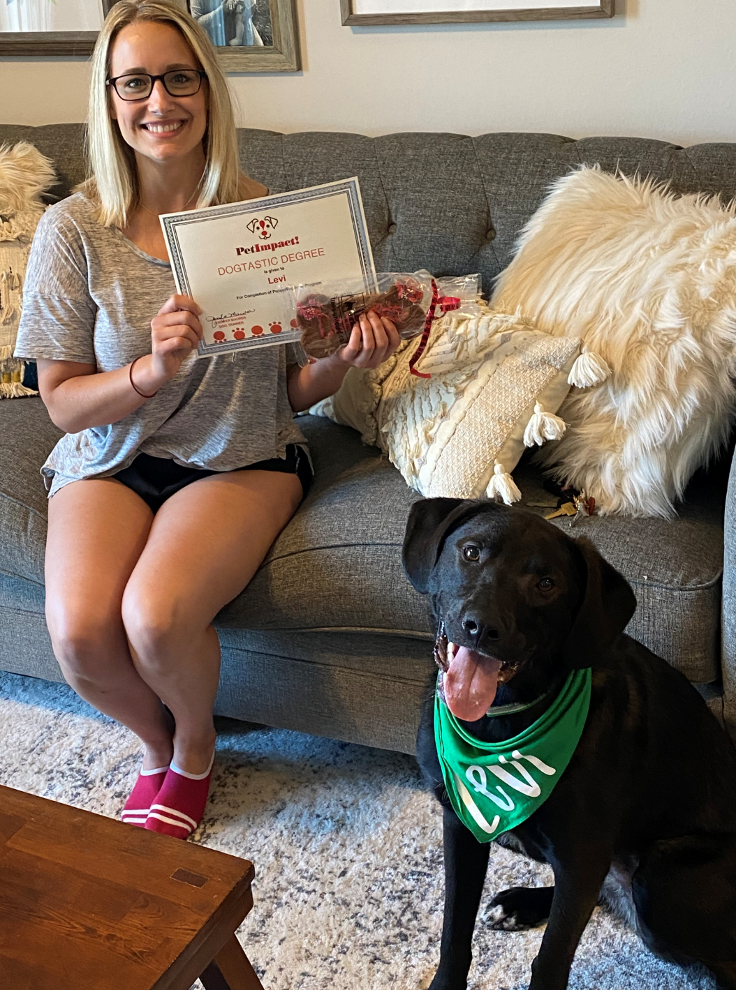 Woman on a couch with a dog; she holds a certificate and treat. Dog wears a green bandana and sits at her feet.