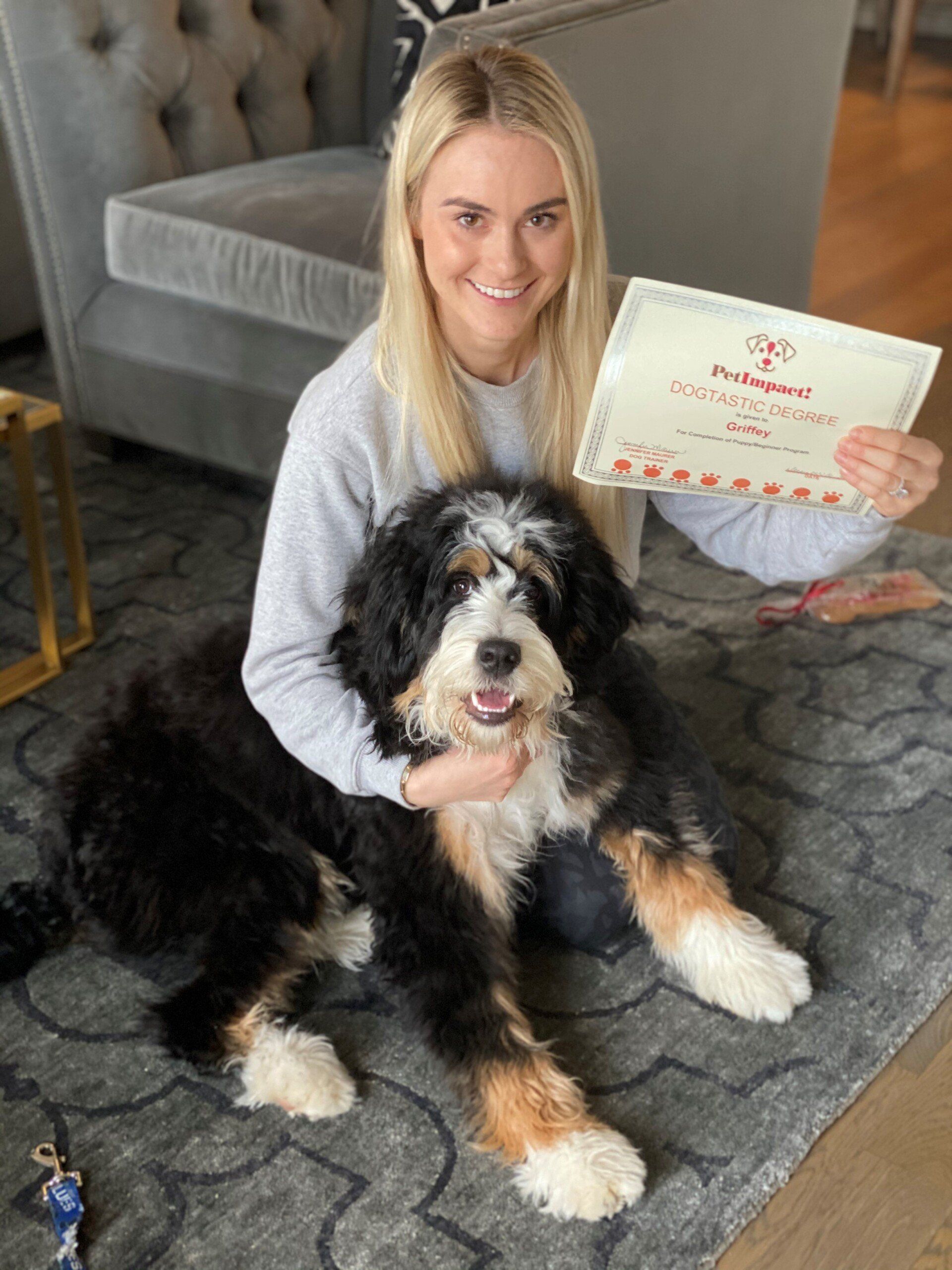 Woman with dog holding a certificate indoors. Dog is black, white, and tan. Both smiling at camera.