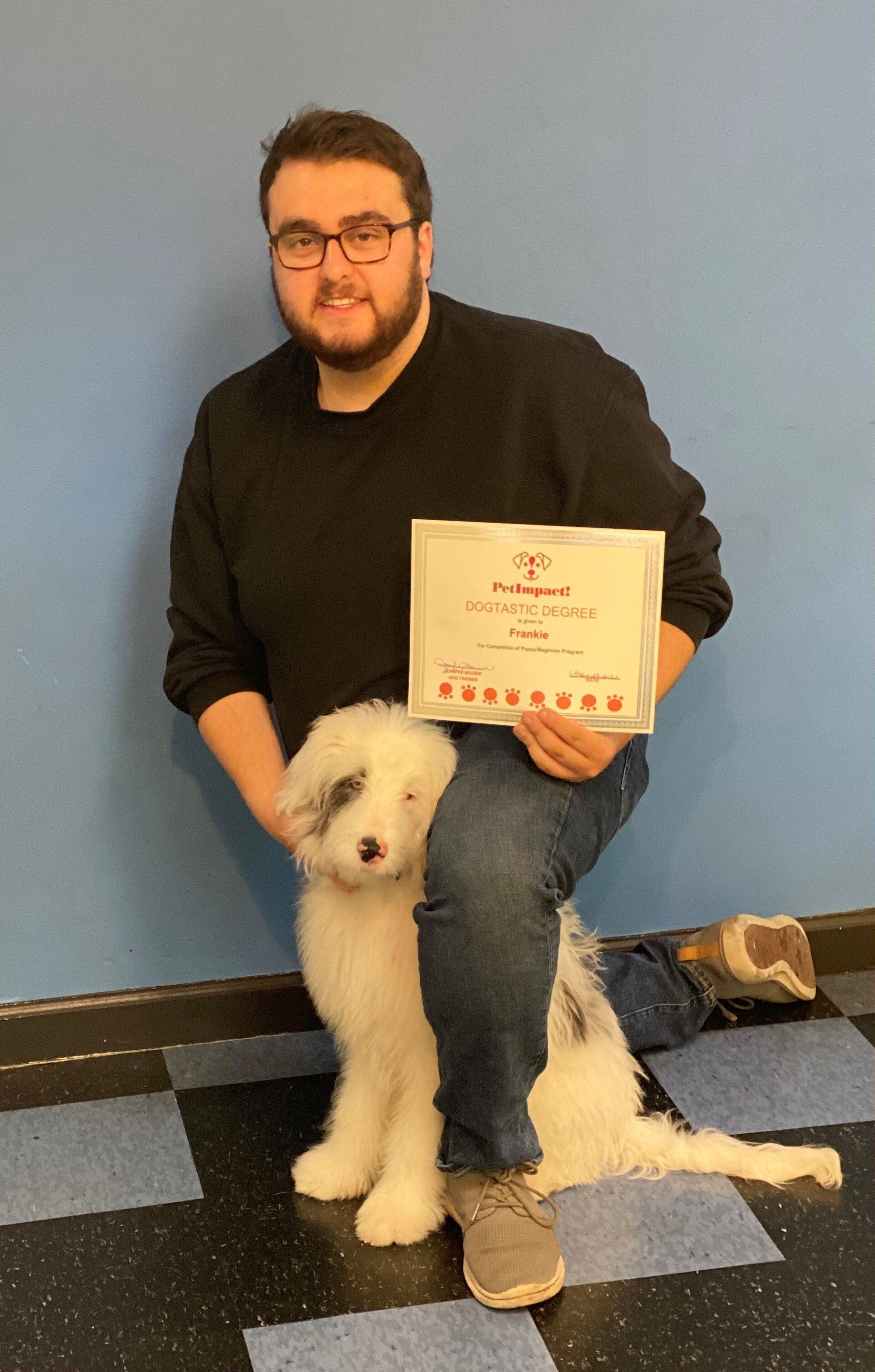 Man and fluffy dog kneeling, holding certificate against blue wall.
