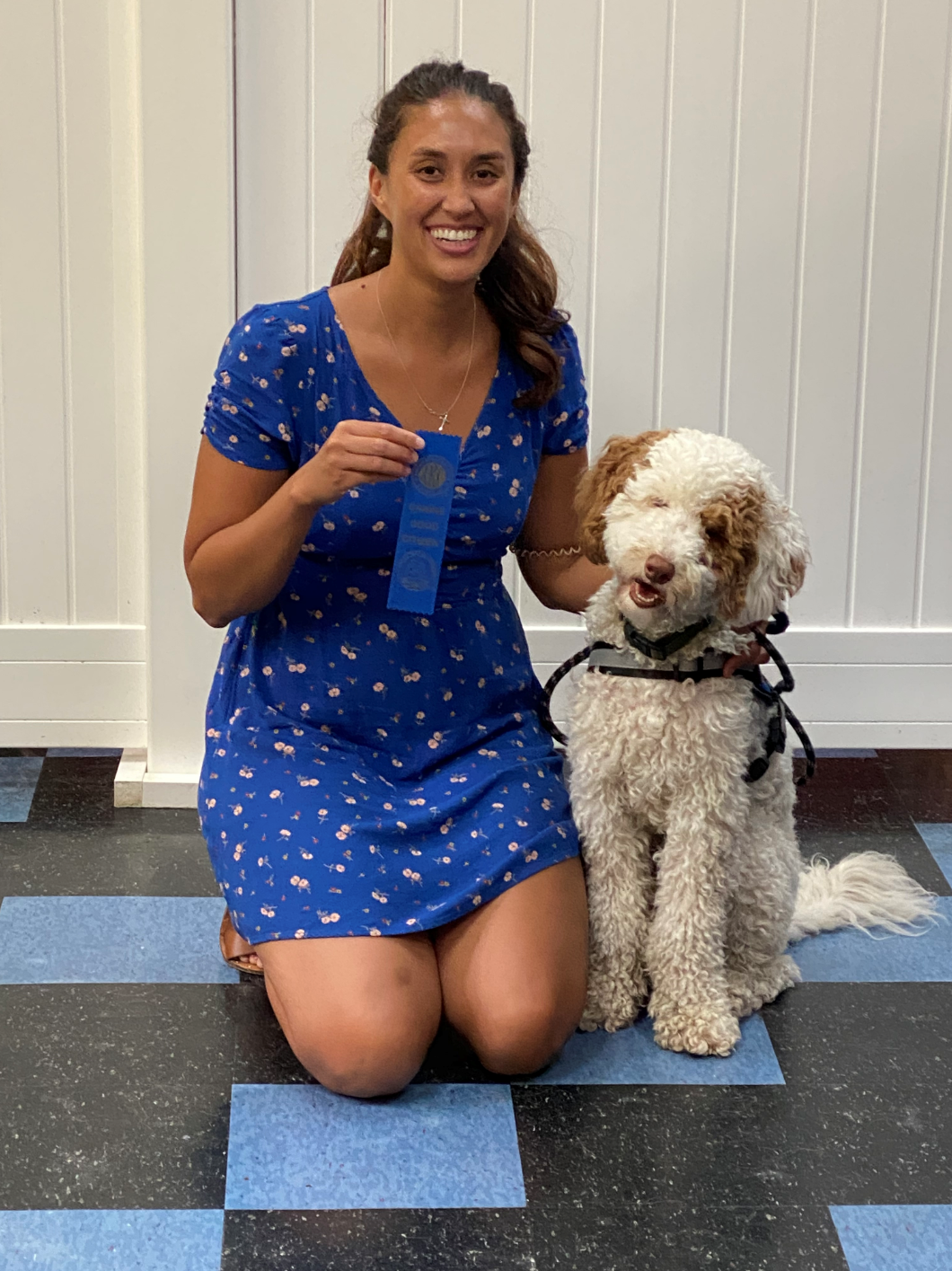 Woman kneels with dog, holding blue ribbon. They are smiling, sitting on a blue and black tiled floor.