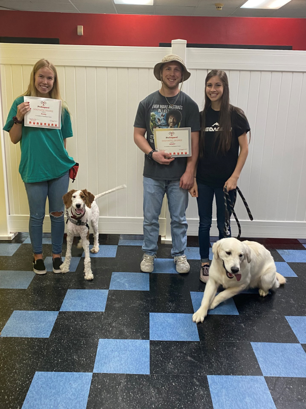 Three people with dogs pose with certificates.  Two people are holding certificates, and all dogs are on leashes in a training room.