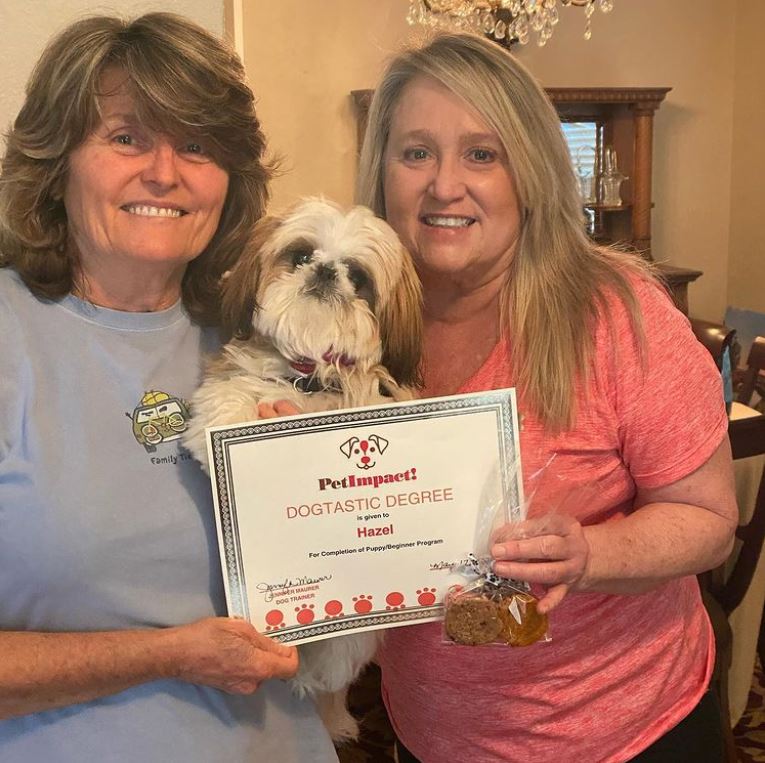 Two women and a dog hold a certificate, presumably for the dog. They are smiling indoors.