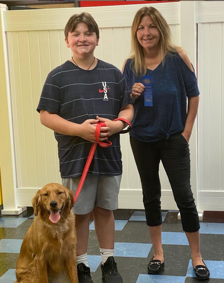 A boy and woman with a golden retriever dog. They hold a red leash, blue ribbon, and smile. Indoors near a white fence.