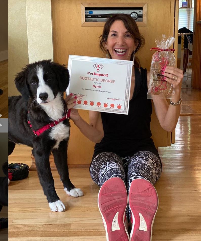 Woman and puppy seated on the floor. Woman holds a certificate and treat bag. Puppy wears a red harness.
