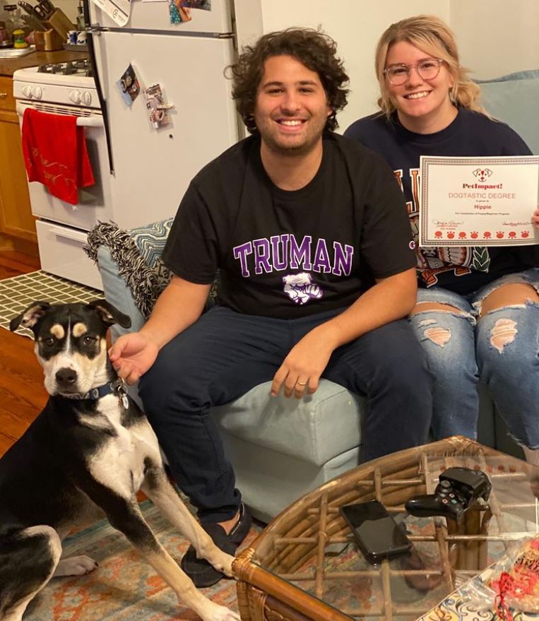 Couple and dog on a couch, holding a certificate. Man wears a Truman shirt.