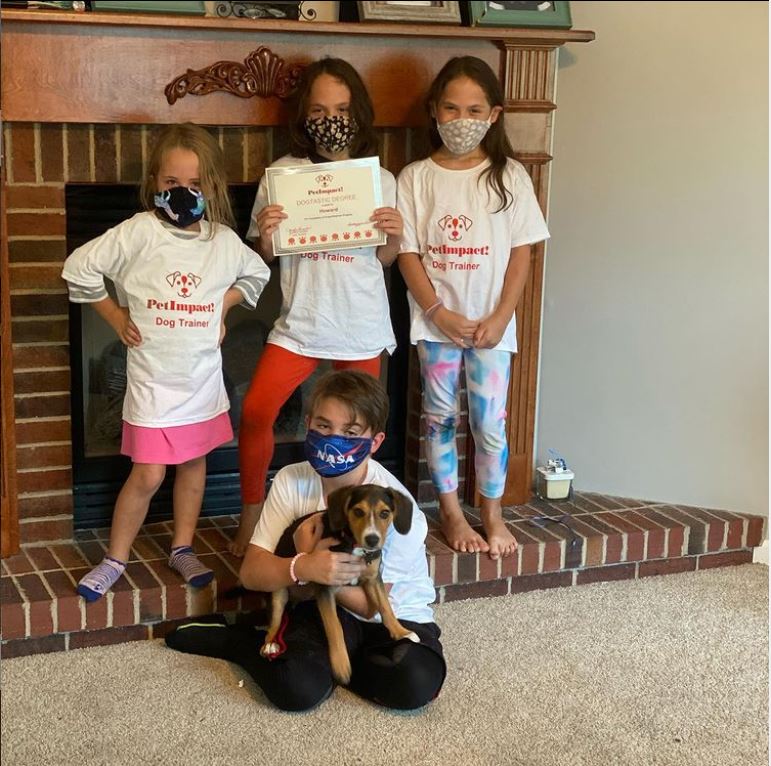 Four children wearing masks and t-shirts pose with a dog in front of a fireplace, holding a certificate.