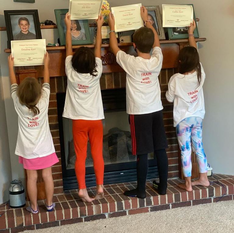 Four children in white shirts holding up certificates above a fireplace.