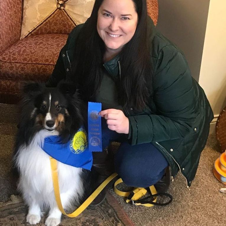 Woman kneels with a dog, holding a blue ribbon. Both wear blue and yellow. Indoors, near a chair.
