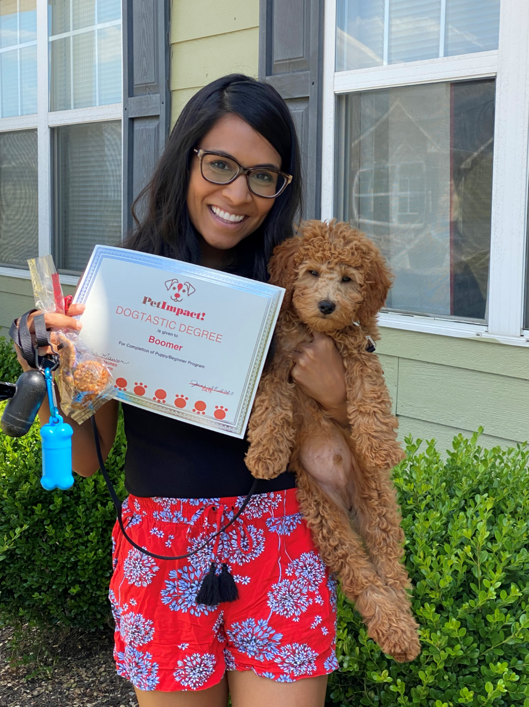 Woman holding a goldendoodle puppy and a certificate, smiling. Outside in front of a house.
