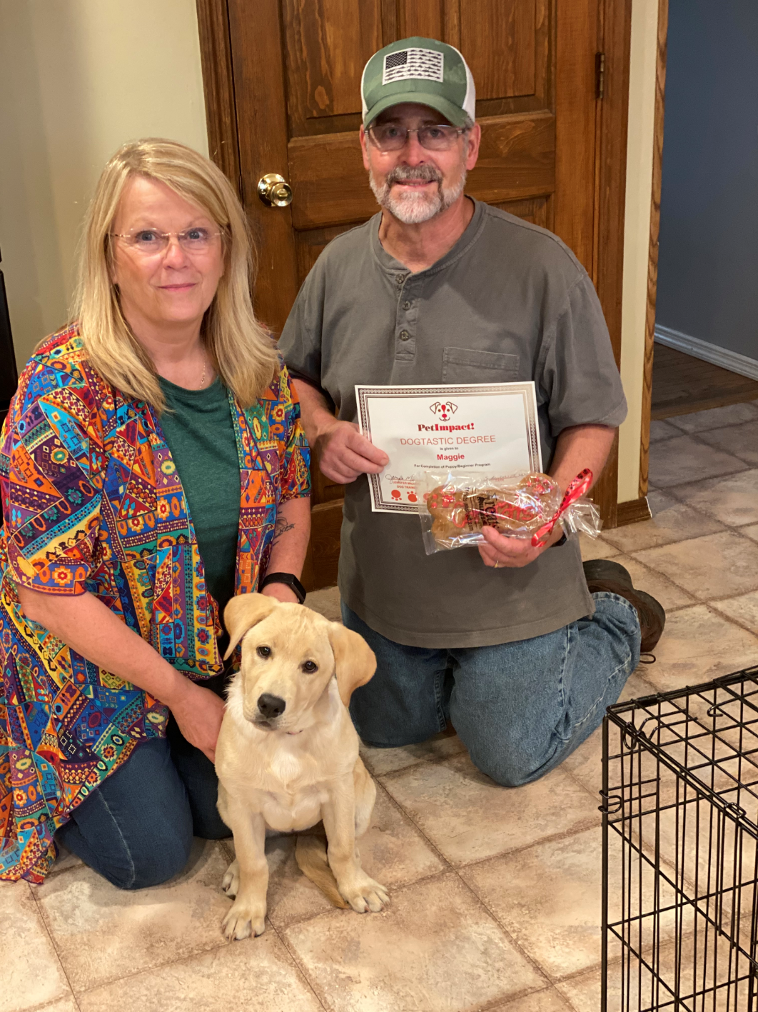Woman and man kneel with a yellow lab puppy. Man holds a certificate and treats; they are indoors.