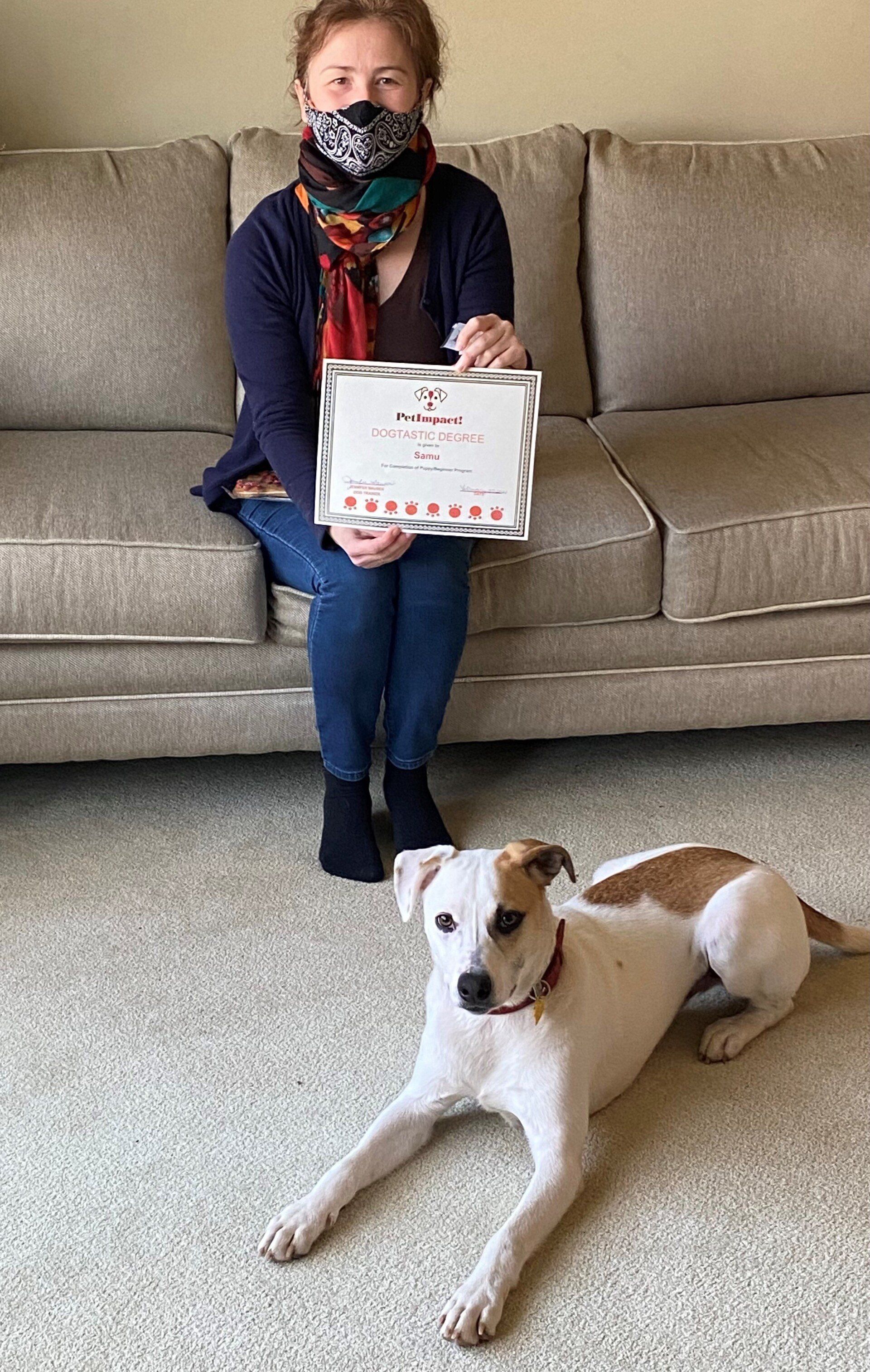 Woman in mask holds framed certificate, seated on a couch; dog lying on the floor in front of her.