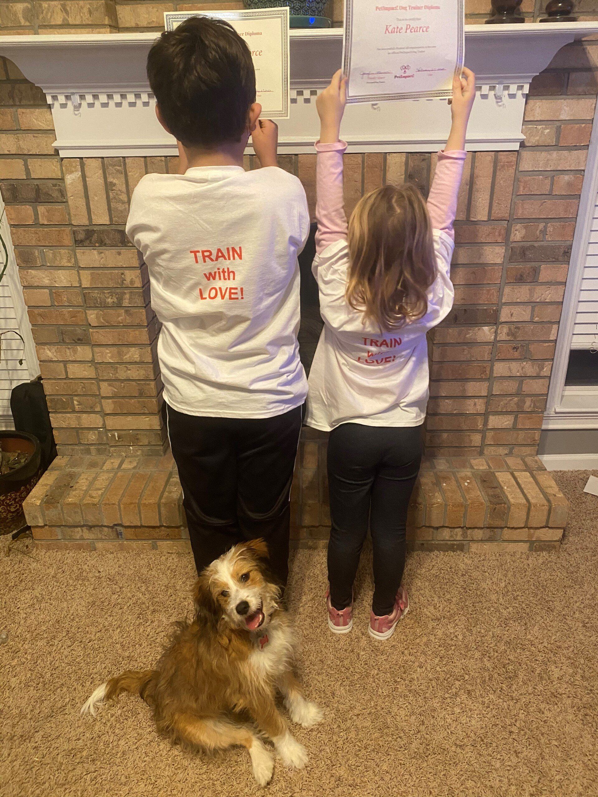 Two children and a dog pose in front of a fireplace, holding a paper. The kids wear shirts that say 