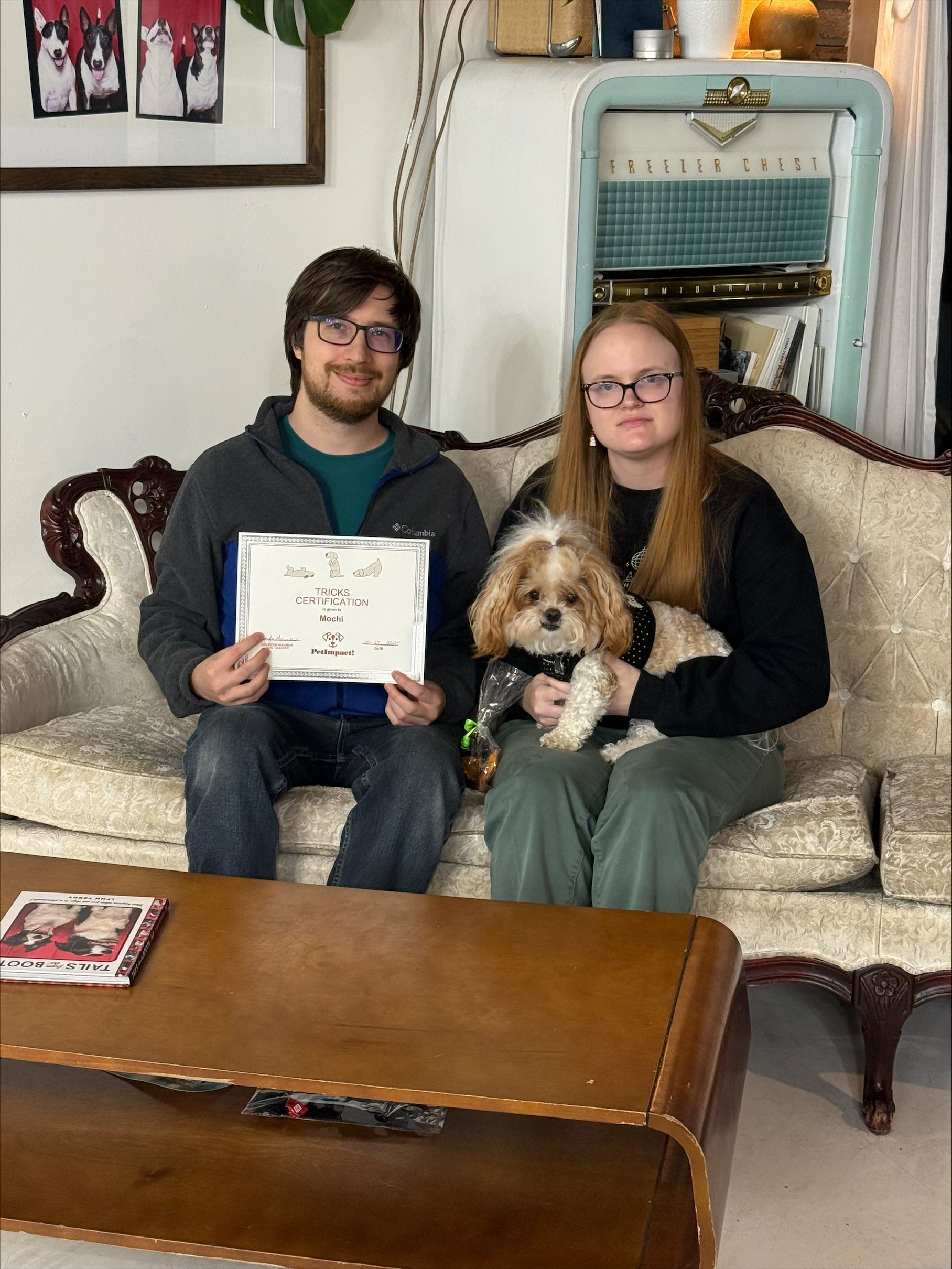 A couple sits on a vintage couch with a dog; the man holds a certificate. Interior setting.