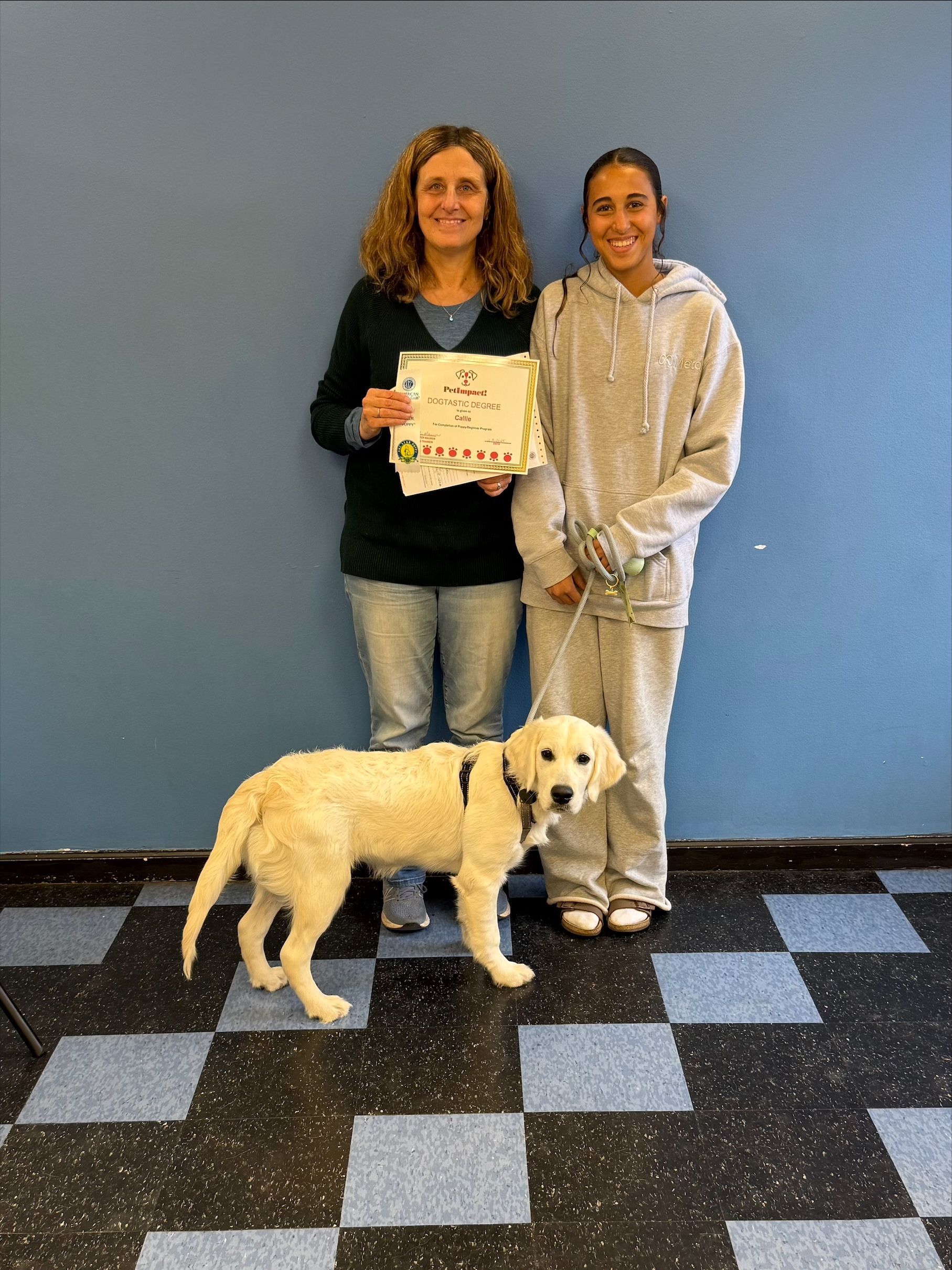Woman and girl with puppy, holding a certificate, standing in front of a blue wall and checkered floor.