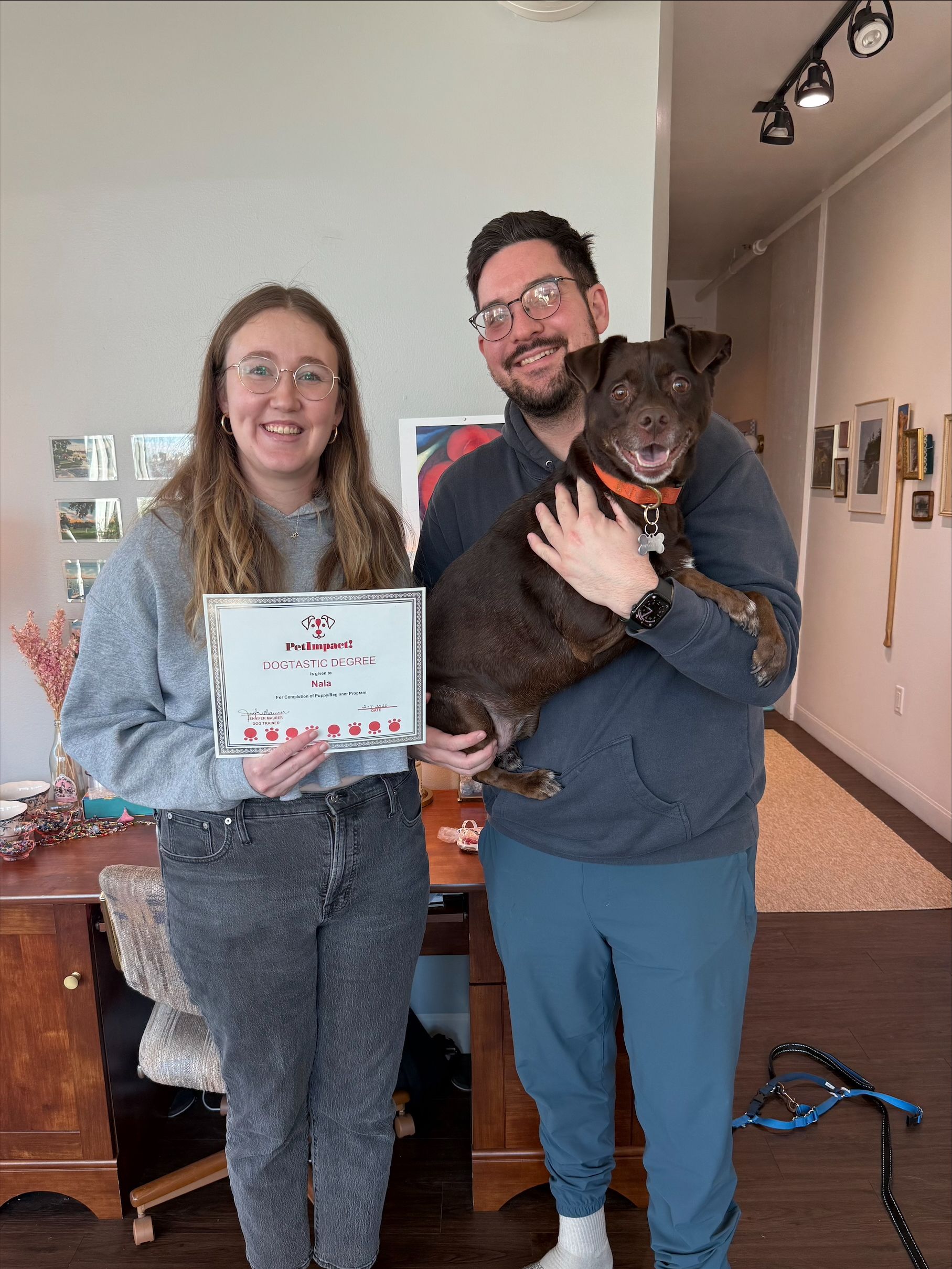 A person holds a sign while standing next to another person who is holding a dark-coated dog in a sunlit room.