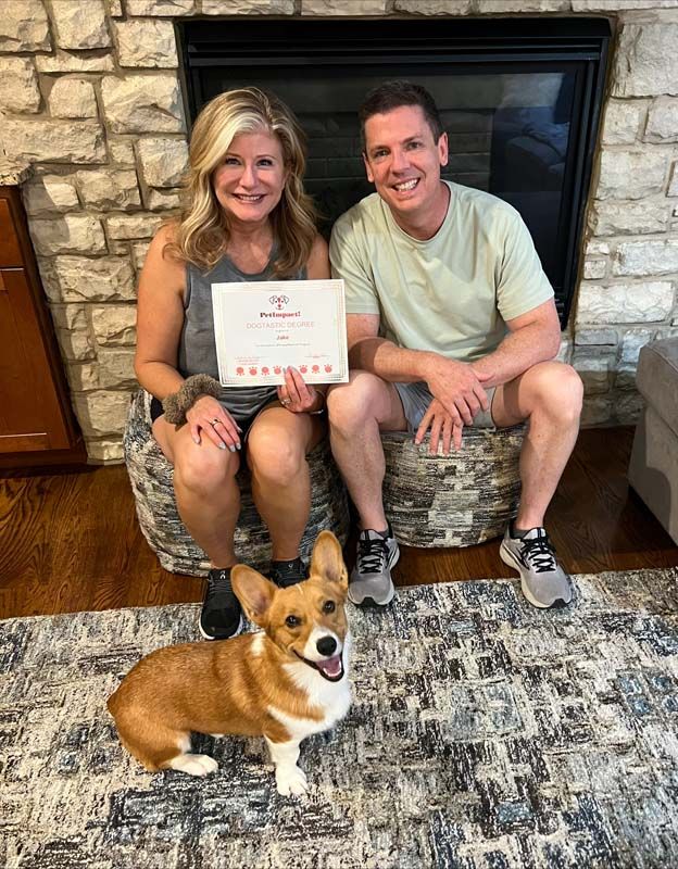 Woman, man, and corgi smile, holding document by fireplace.