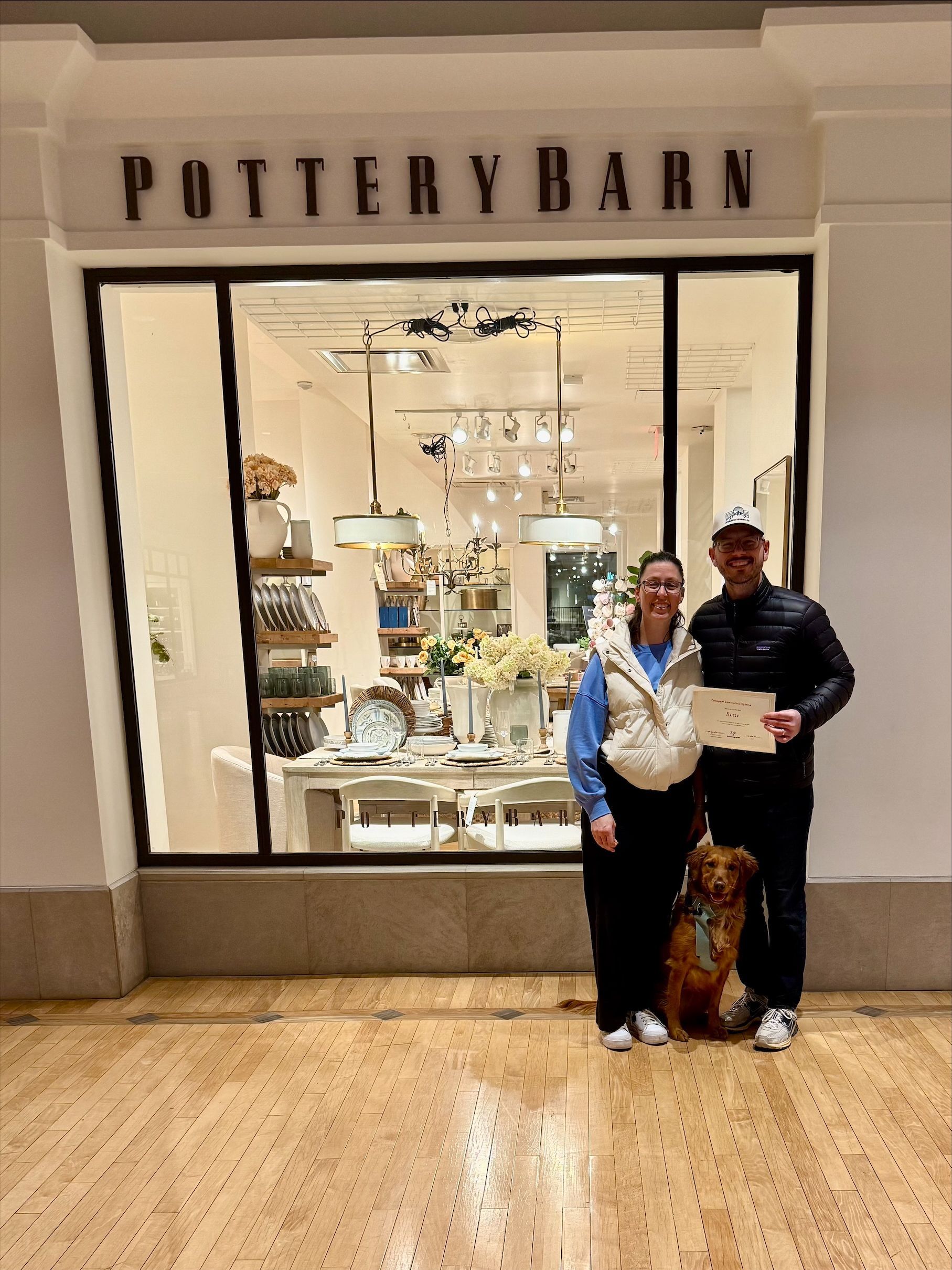 Couple and dog pose in front of a Pottery Barn store. Man holds a paper.