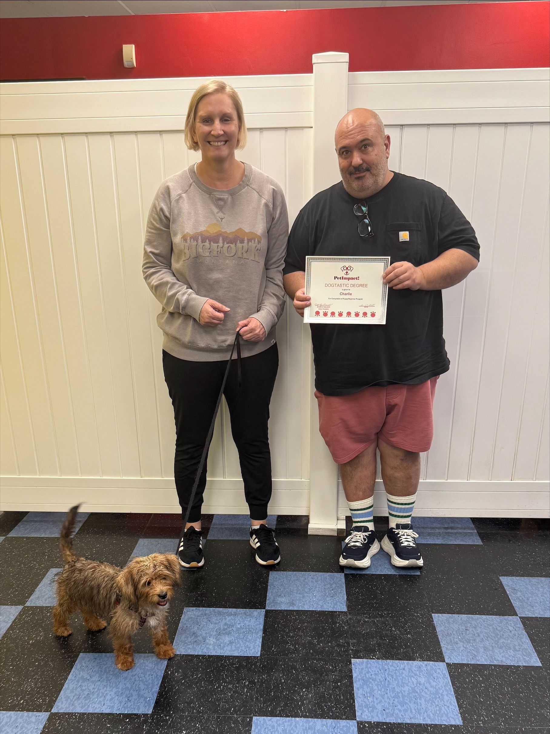 Woman holding a leash, man holding a certificate, small dog. Standing in front of a white fence and checkered floor.