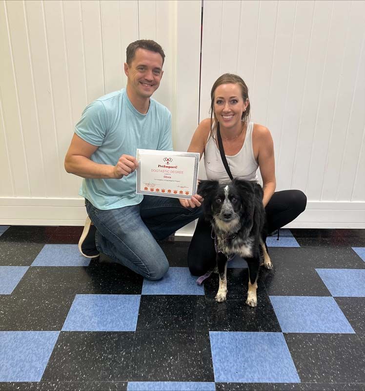 Couple and dog pose with a certificate in a room with a checkered floor.