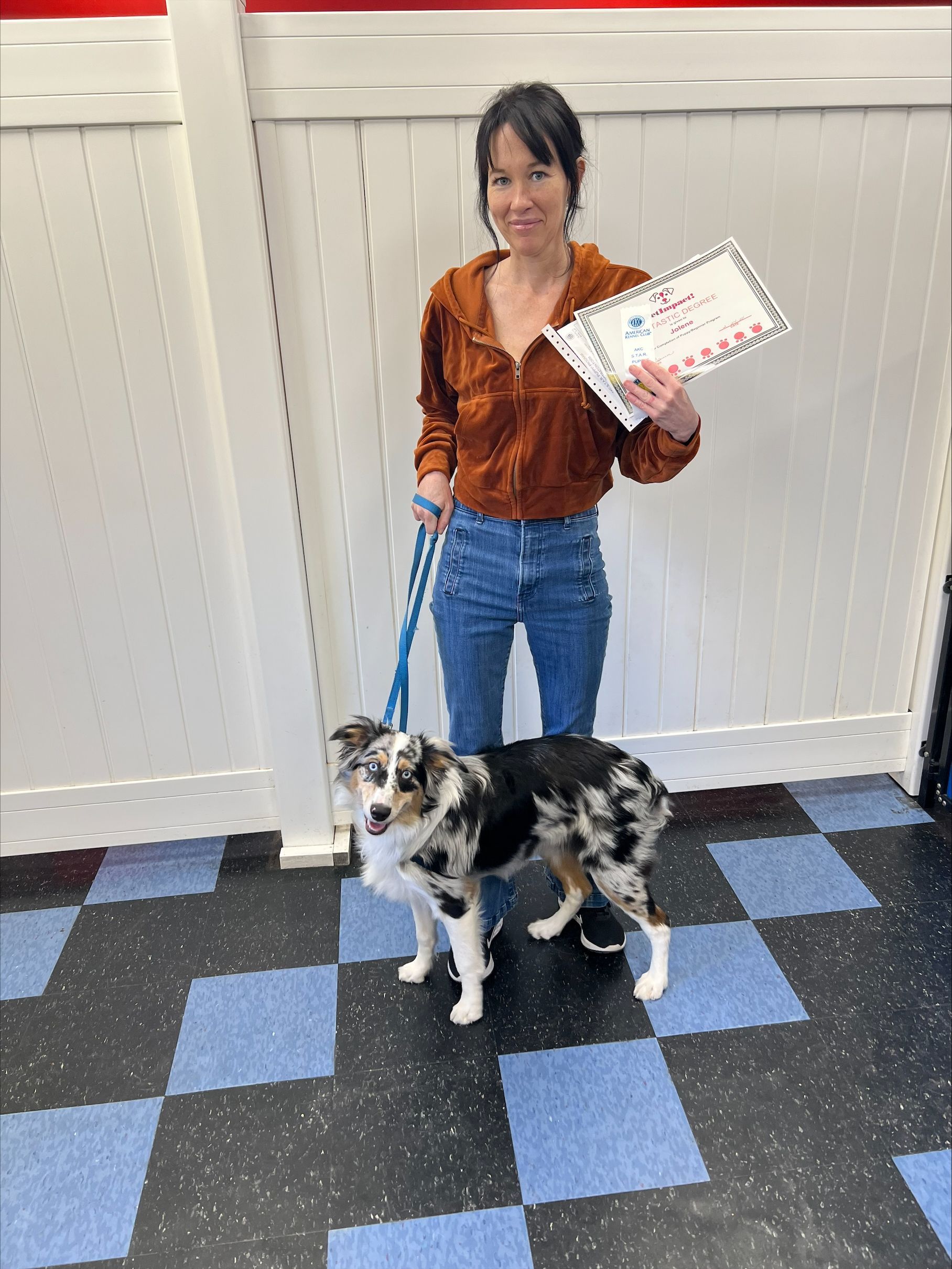 Woman with dog, holding a certificate. She wears a brown jacket and jeans. Dog is black, white, and brown. Standing on checkered floor.