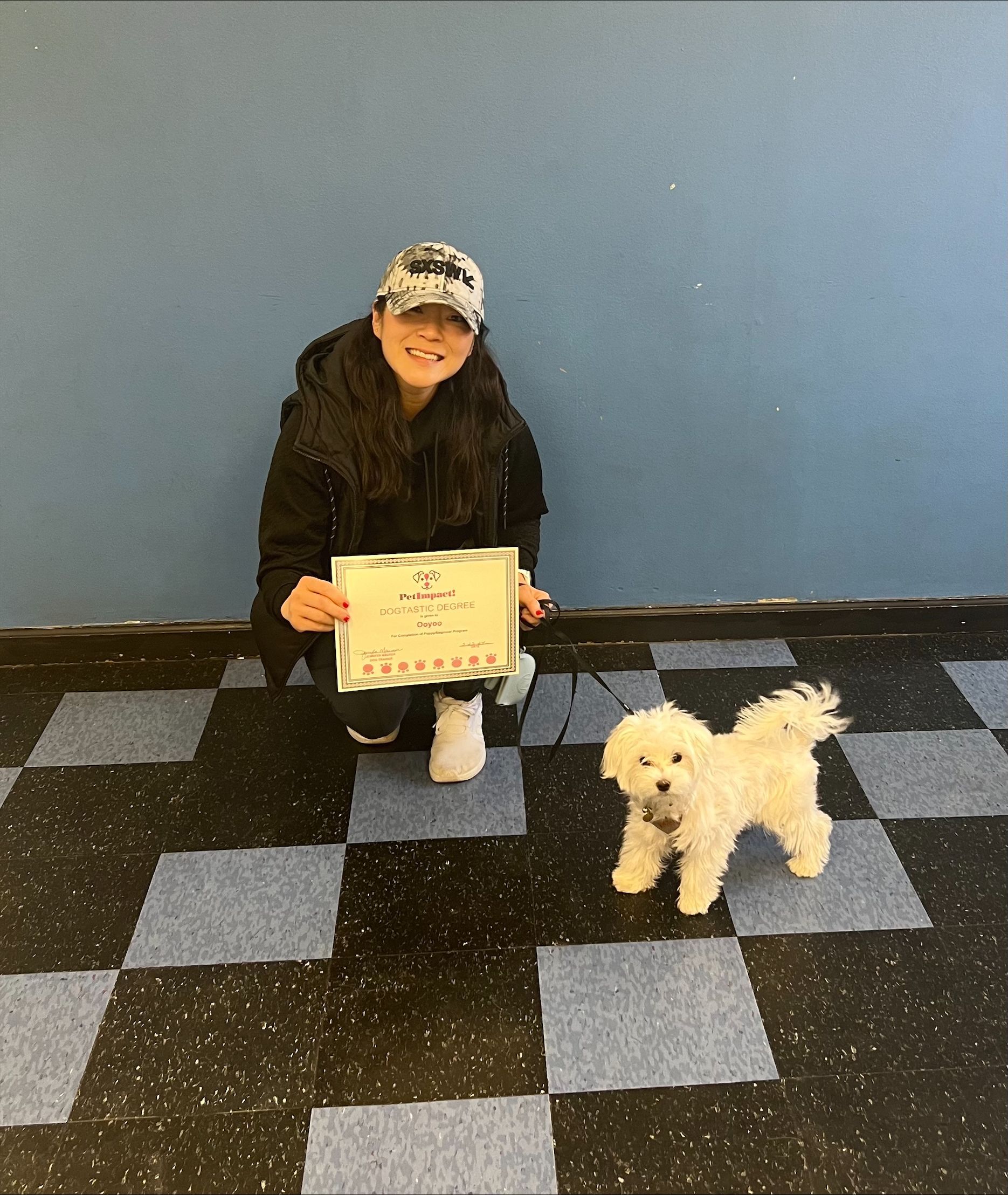 A woman kneeling next to a small white dog holding a certificate