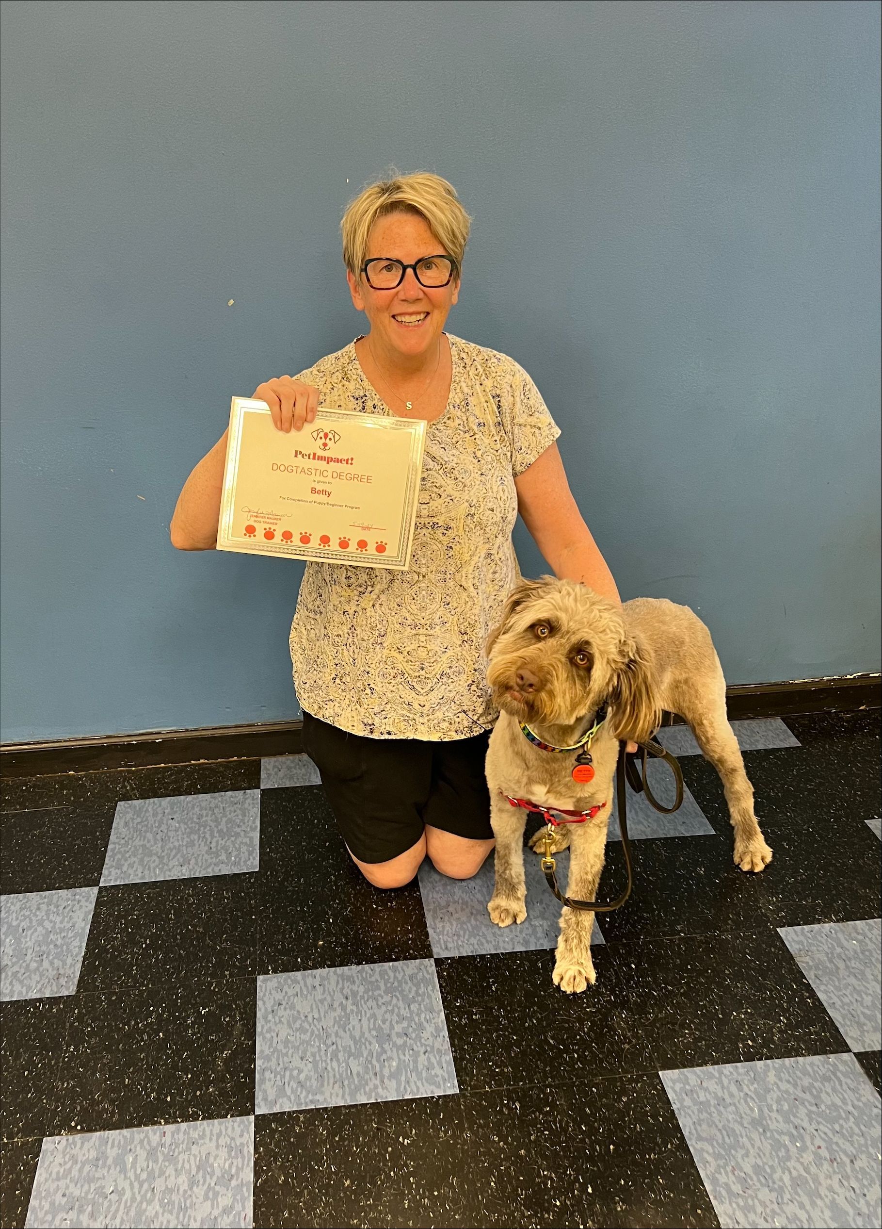 Woman kneeling next to dog, holding paper. Both smiling. Blue wall background, checkered floor.