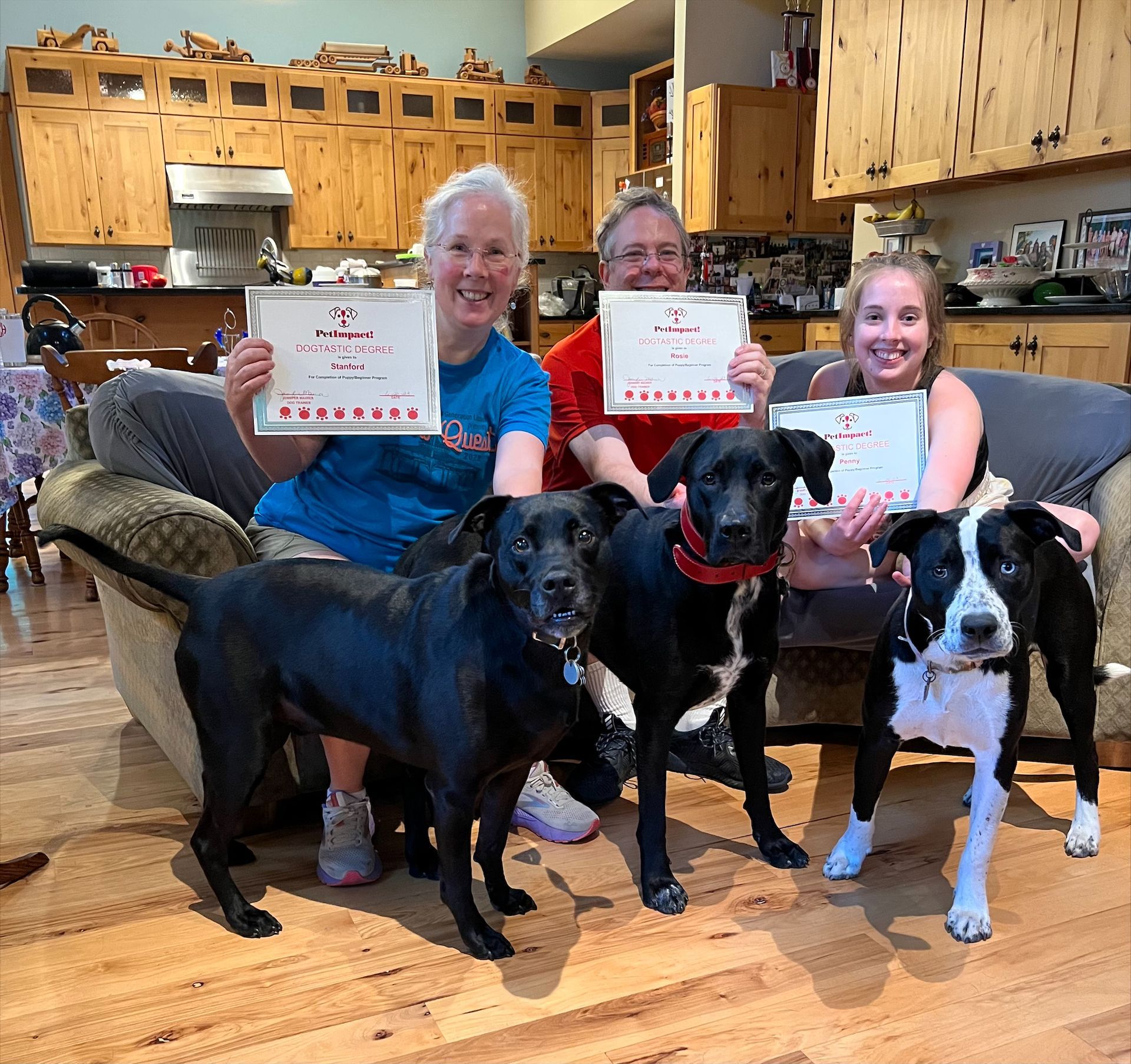Three people and three dogs pose with certificates in a living room with wooden cabinets.