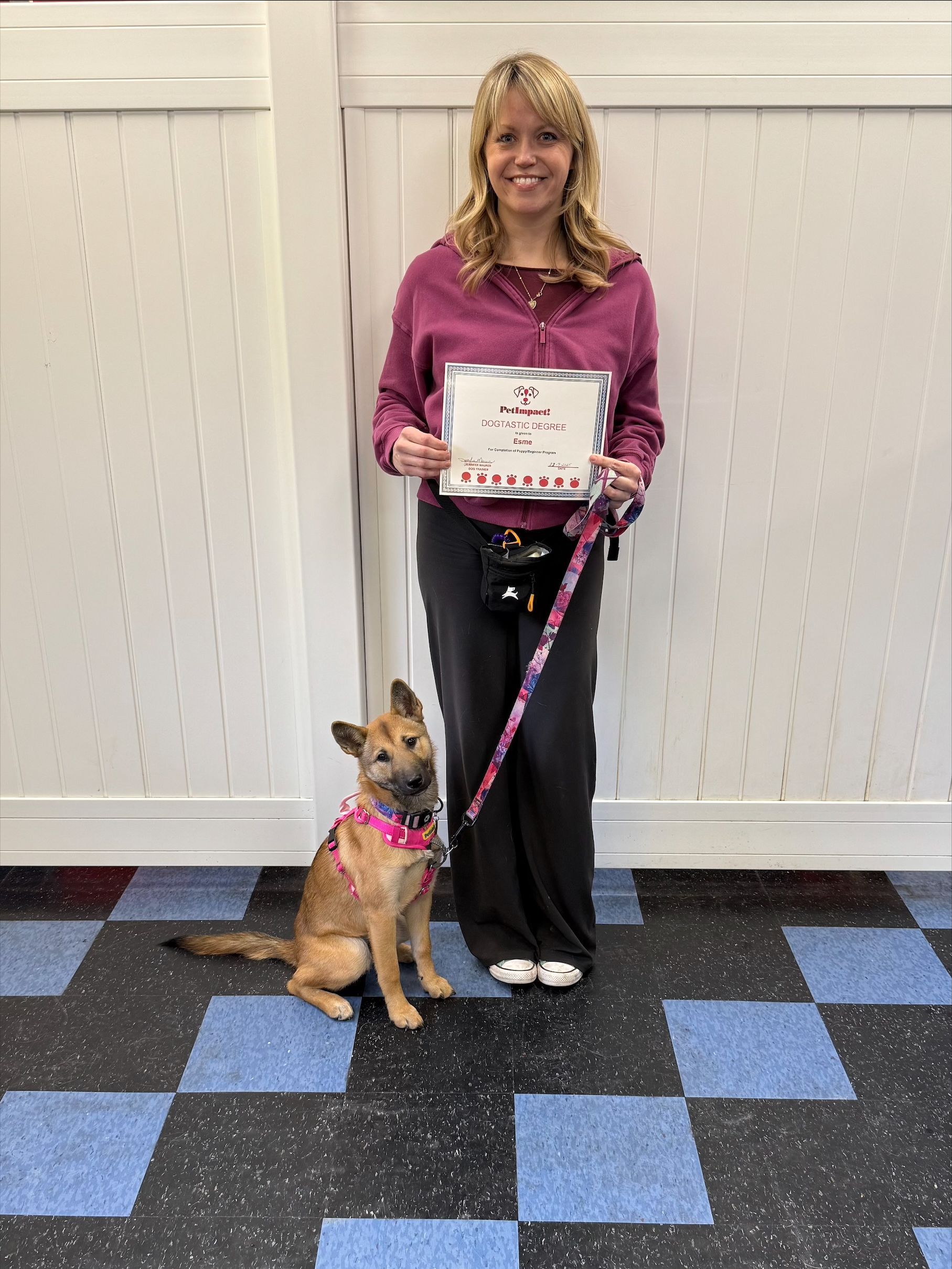 Woman and dog pose, holding a certificate. The dog sits, wearing a pink collar. They are in a room with blue and black checkered flooring.