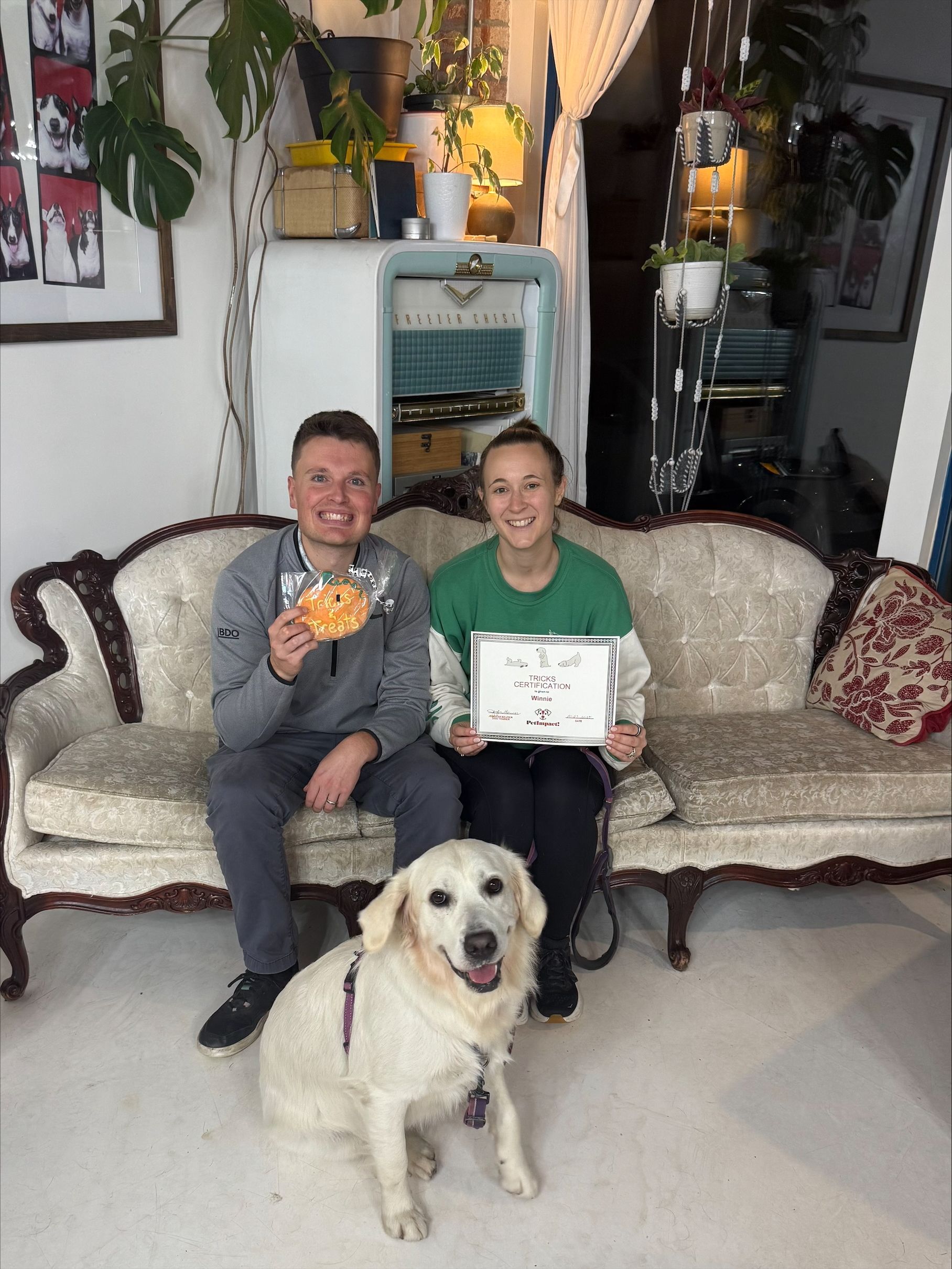 Two people and a golden retriever seated on a vintage sofa. One person holds a donut, the other a certificate. Interior.