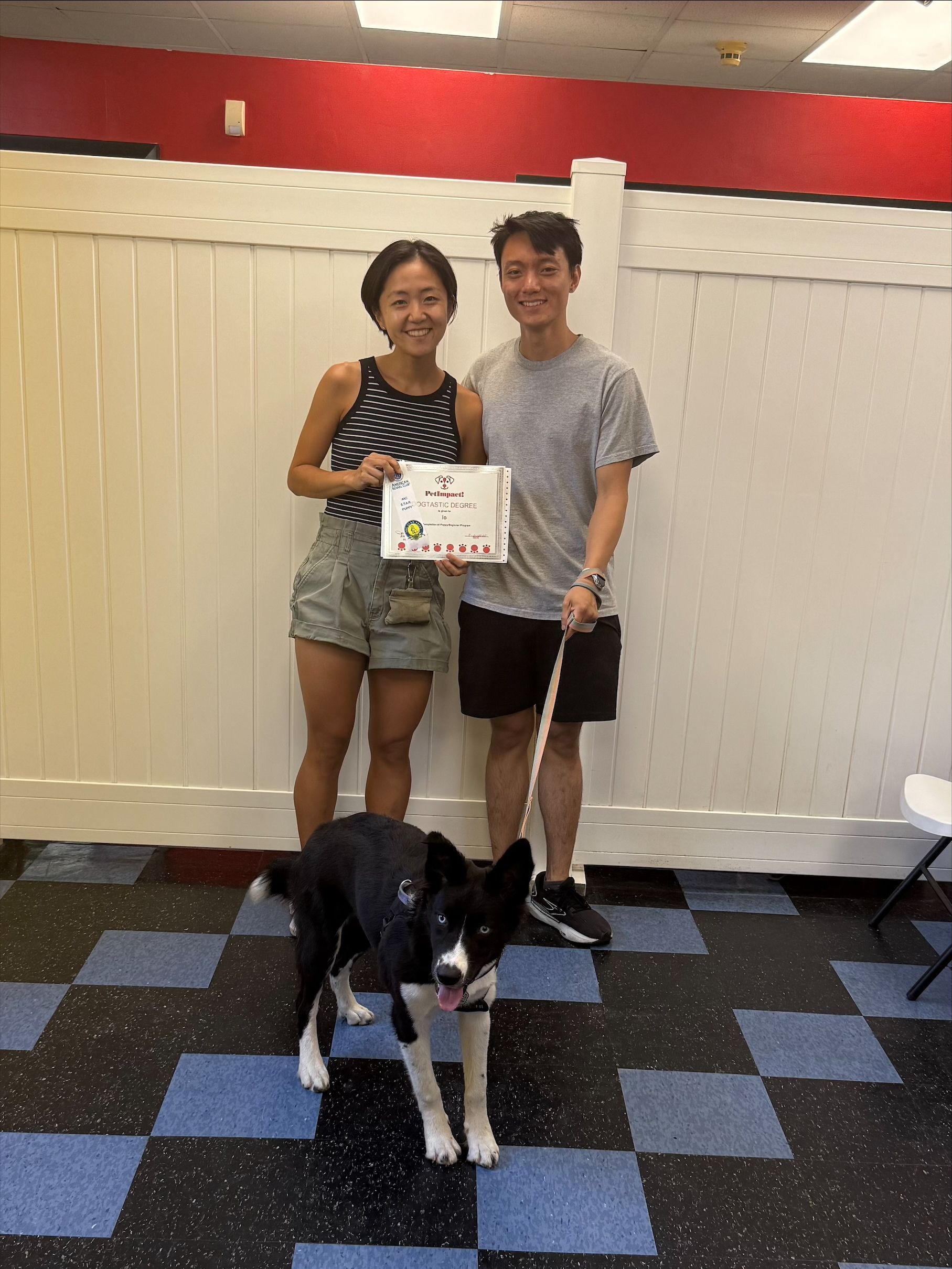 Two people and a dog holding a certificate. The people stand indoors by a fence, smiling. The dog is black and white, wearing a harness.
