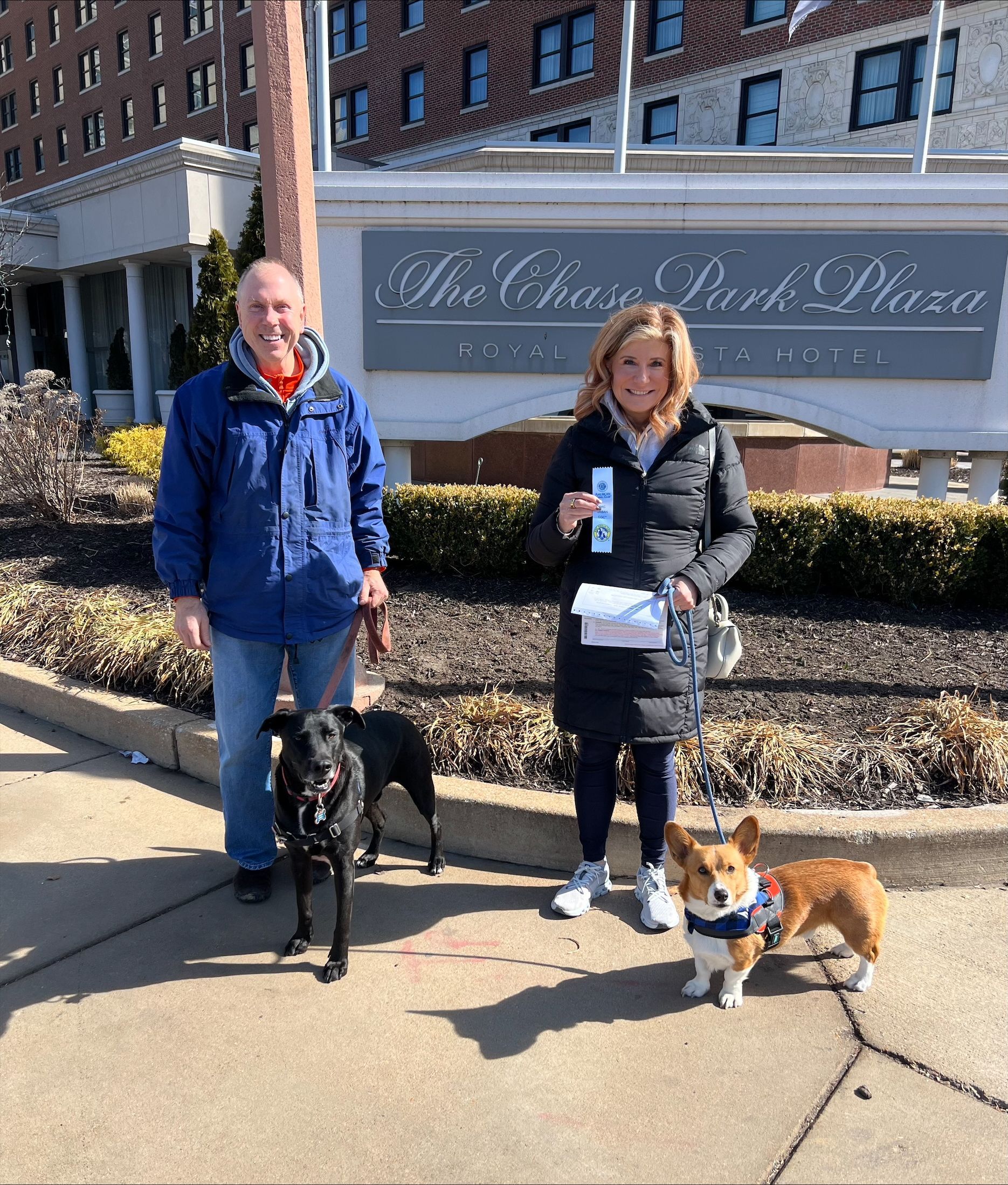 A man and a woman are standing next to two dogs on a sidewalk in front of a building.