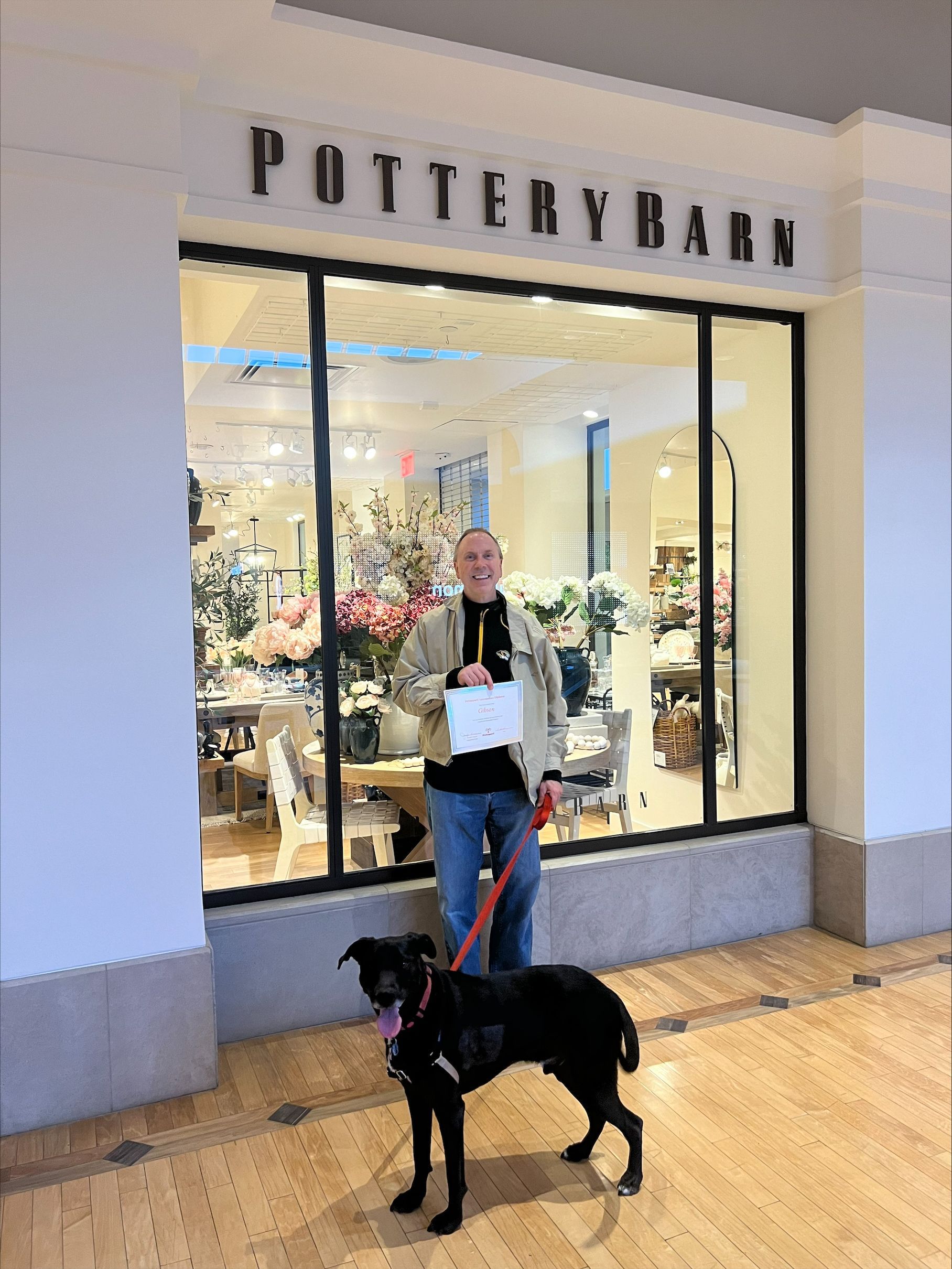 Man with dog outside Pottery Barn store. Man holds paper, smiling. Black dog on leash.
