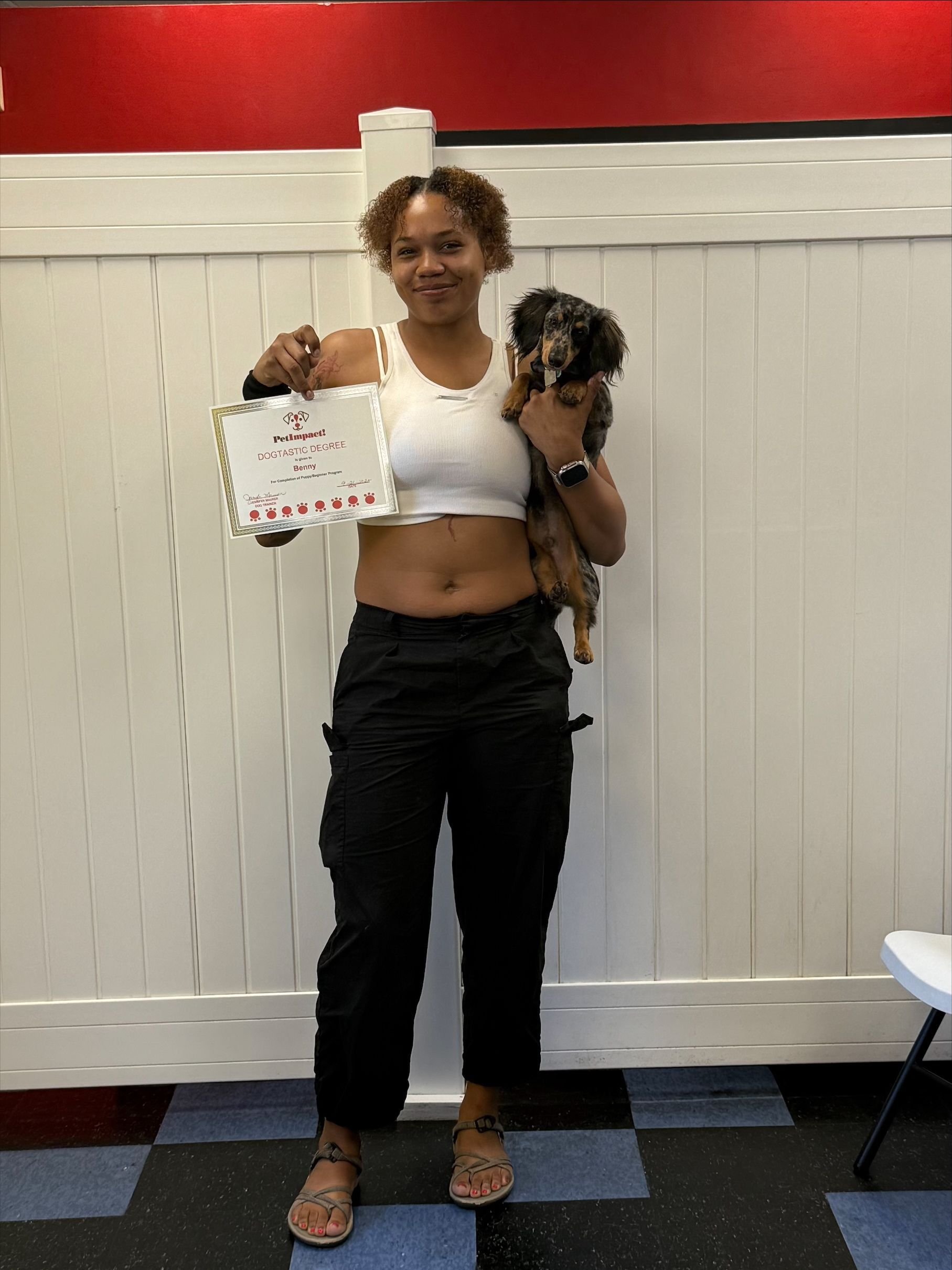 Woman holding a dog and sign. She wears a crop top, black pants, sandals, and smiles. White fence and red wall in the background.